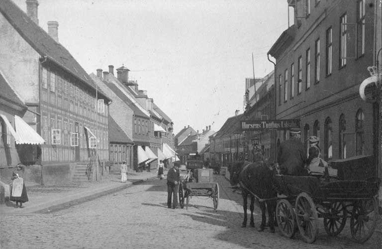 Hestevogn på en brostensbelagt gade omkranset af bygninger og fodgængere, sort-hvid historisk scene