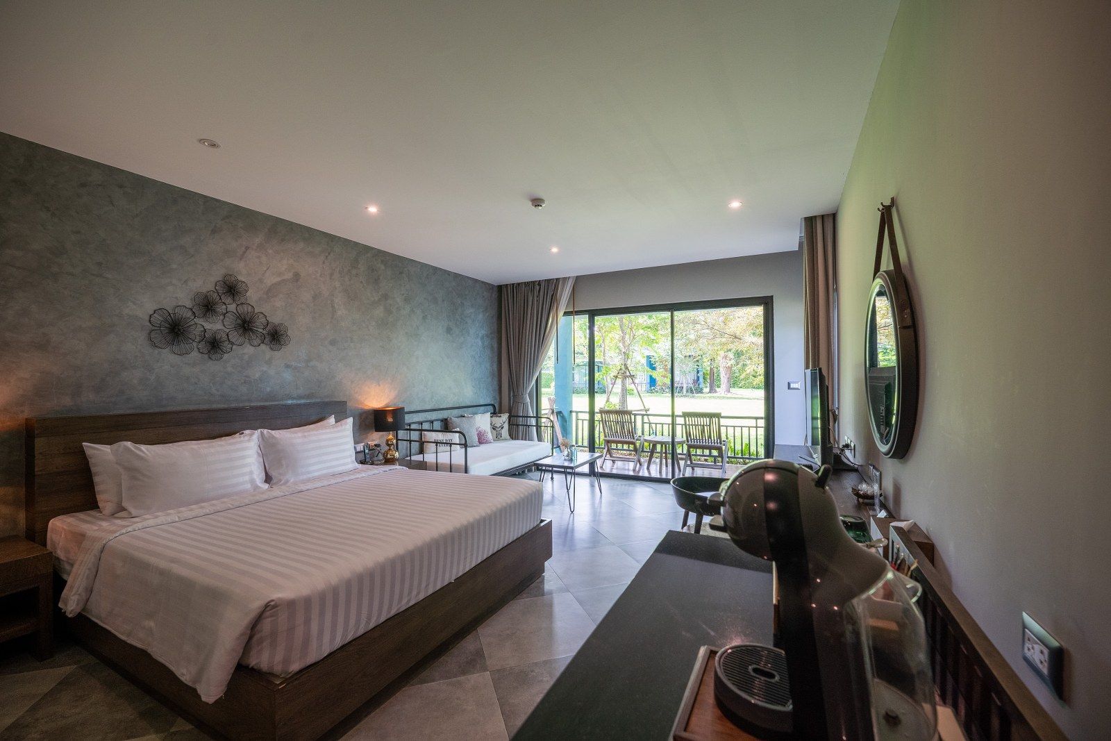 Hotel room with a bed, desk, and sliding door to a balcony overlooking a garden.