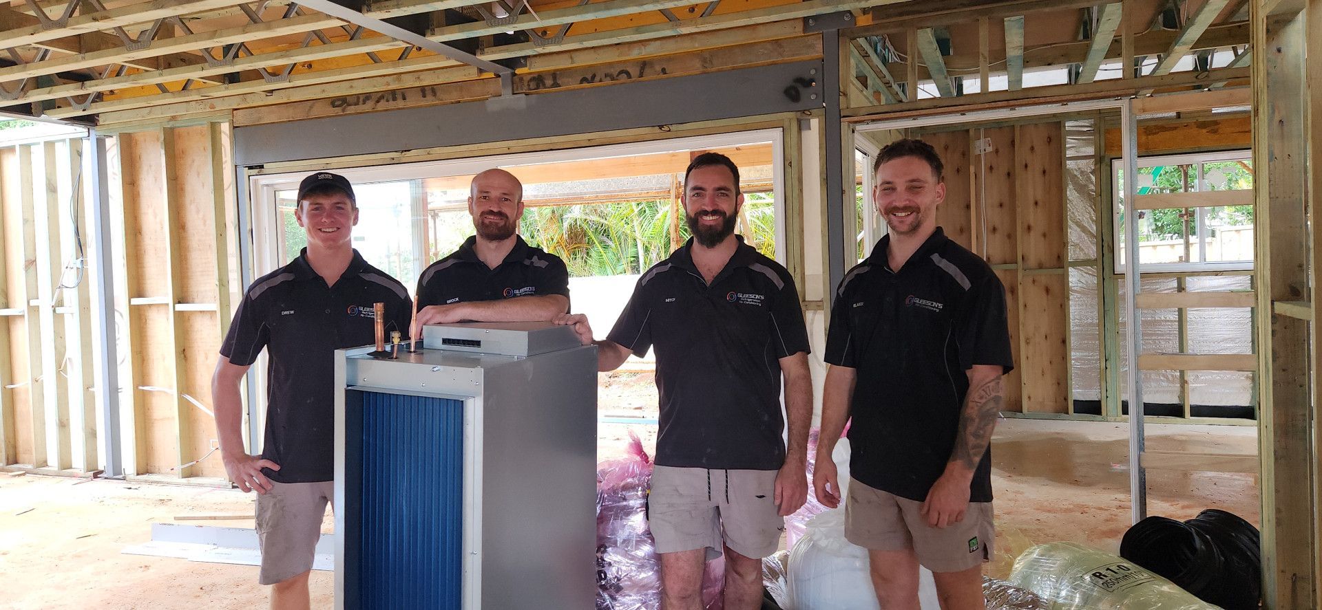 Four men in work uniforms stand in a partially constructed building, posing with a large HVAC unit — Gleeson's Air Conditioning in Port Macquarie, NSW