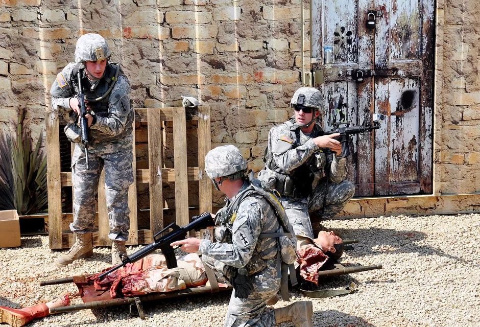 Soldiers in camouflage training with rifles beside a wall and a practice casualty on the ground