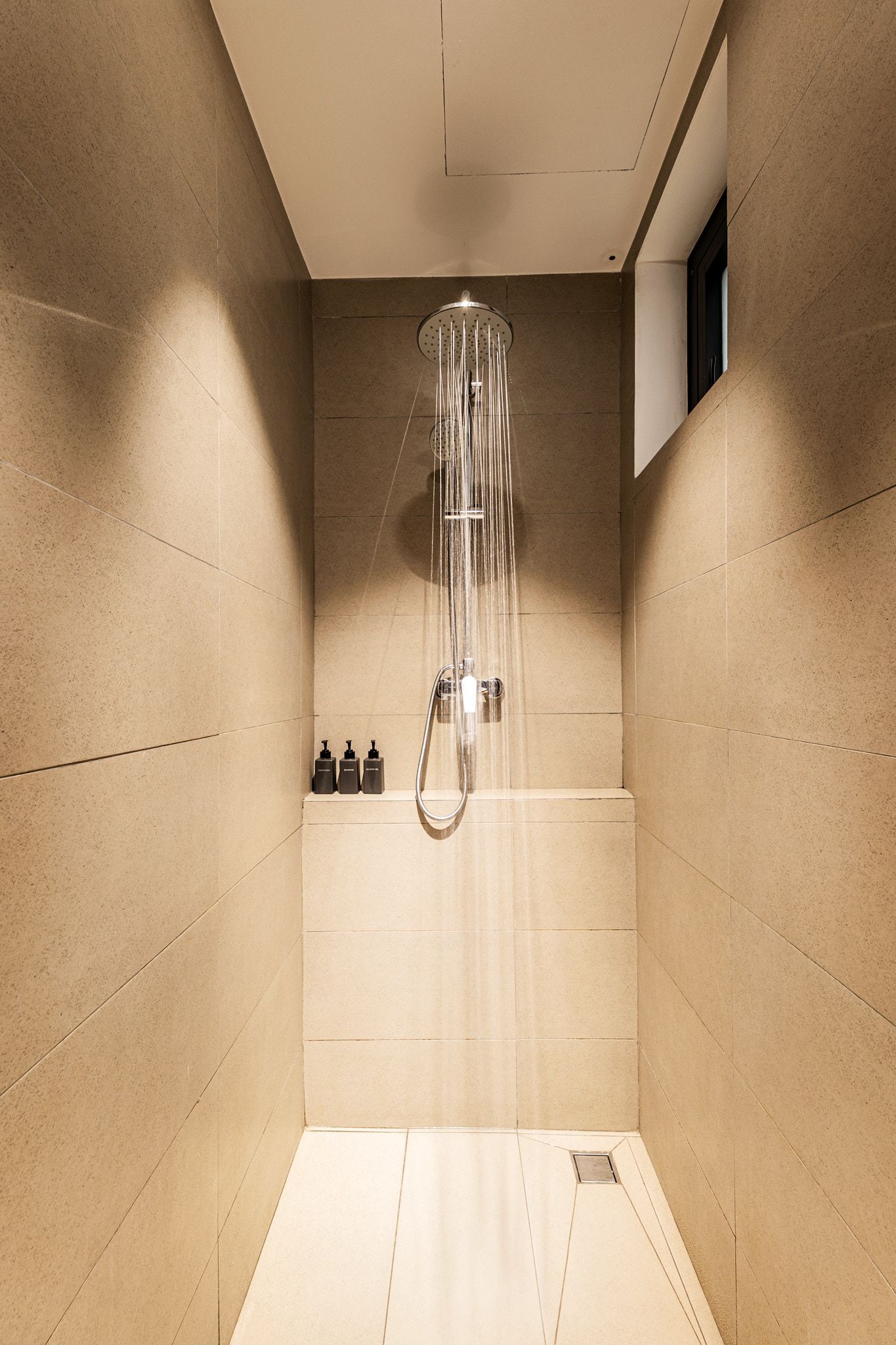 Narrow beige shower stall with a wall-mounted showerhead and small ledge, seen from the doorway