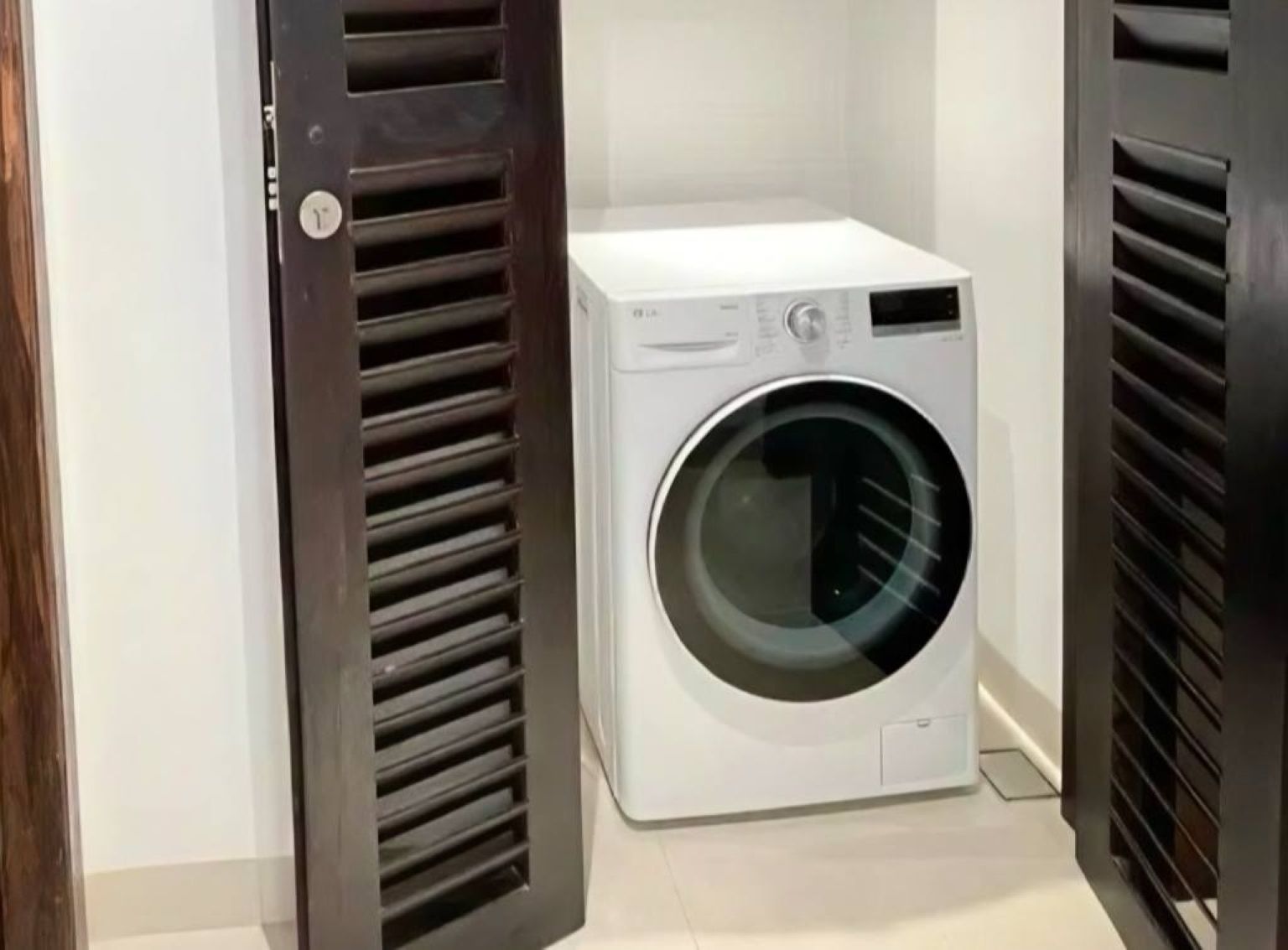 White front-loading washing machine between two dark louvered doors in a narrow utility area