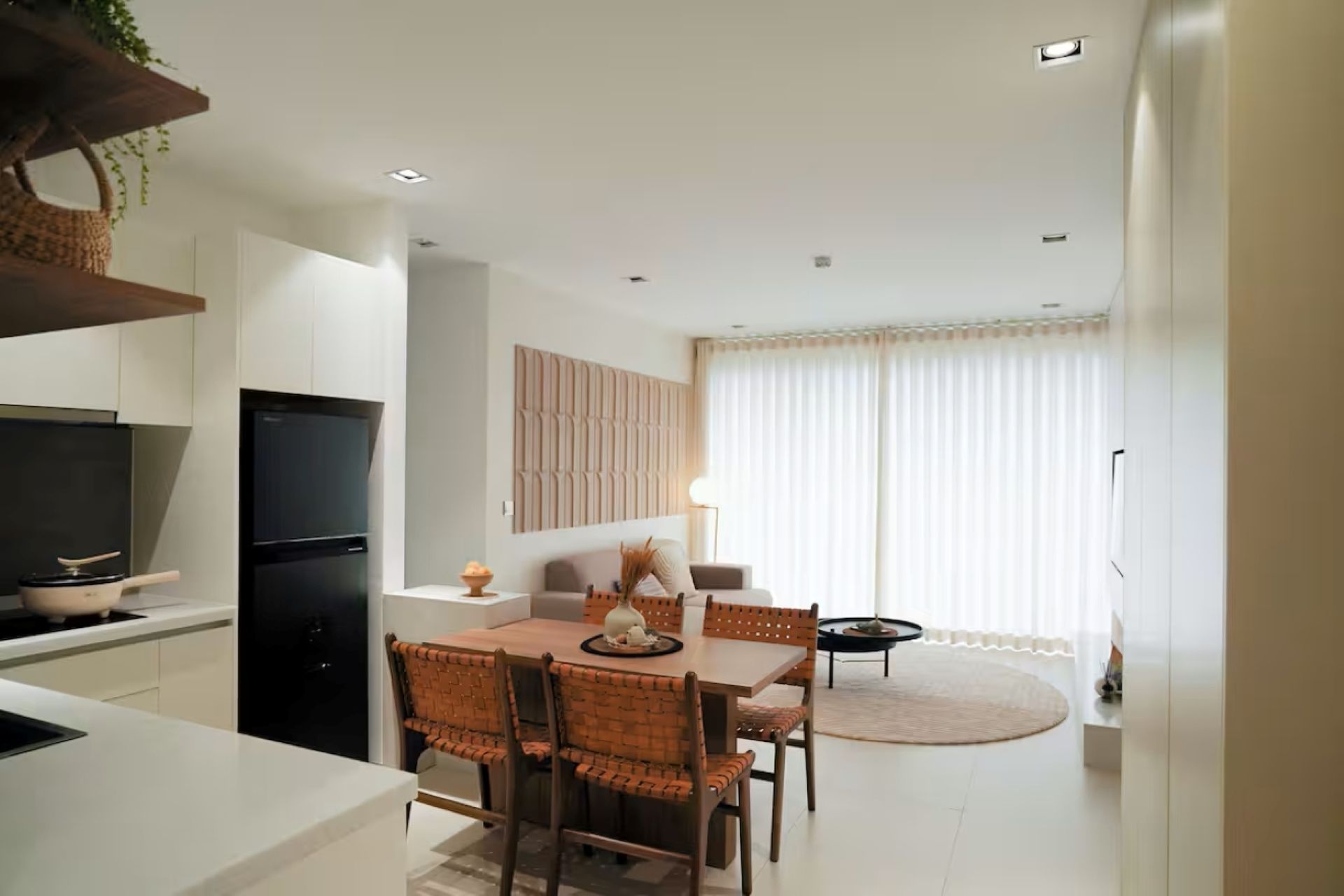 Bright modern kitchen-dining room with wooden table, woven chairs, black fridge, and sheer curtains.