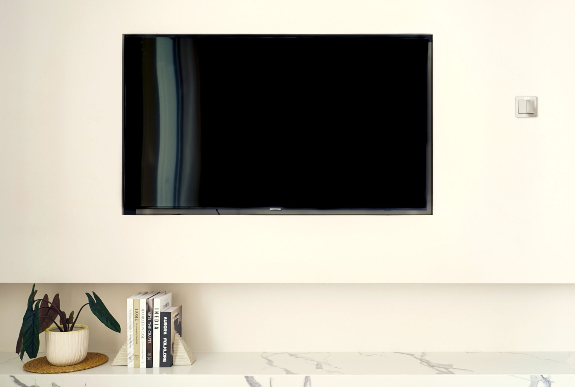 Wall-mounted black TV above a shelf with books, decor, and a white countertop in a minimal room