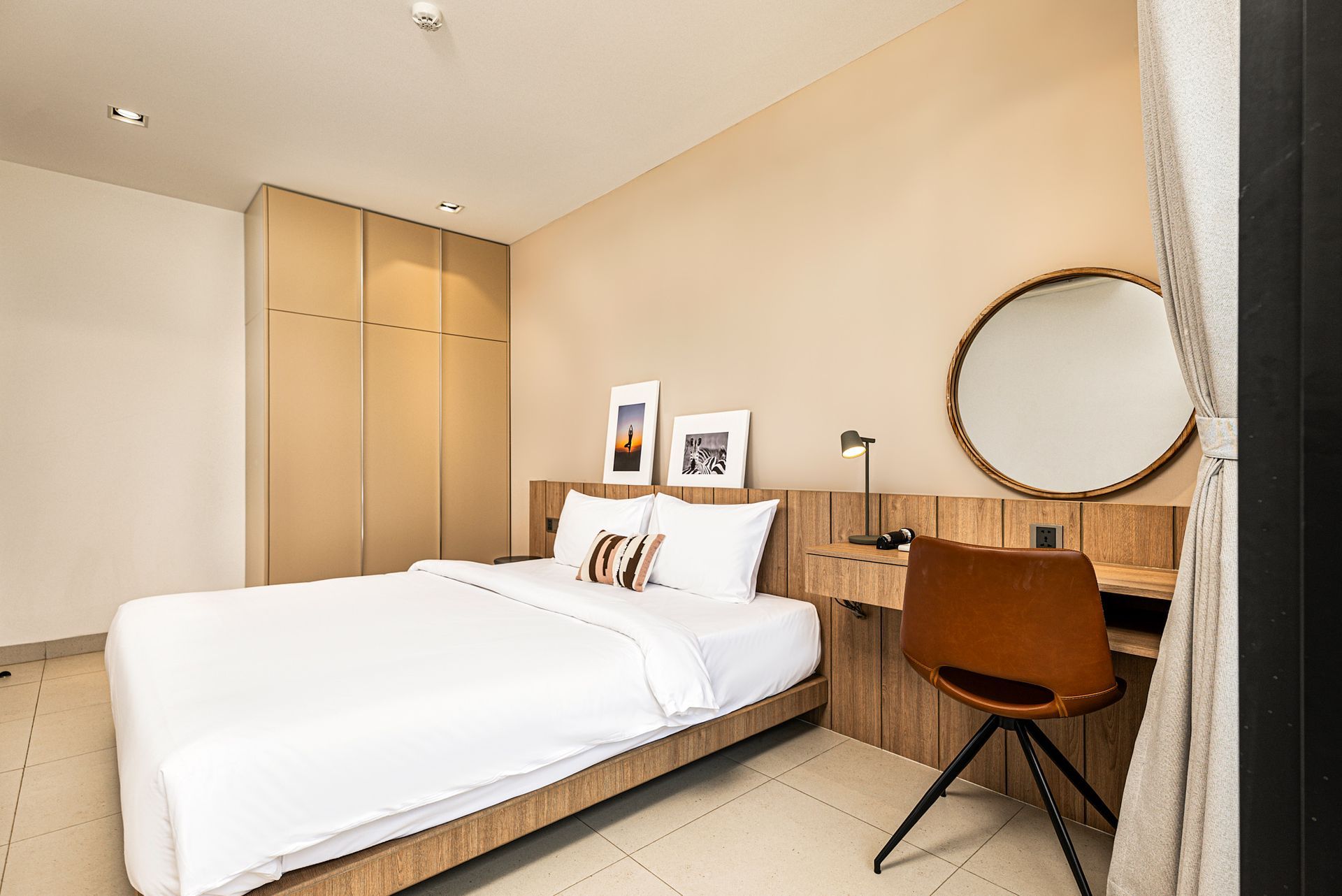 Minimal bedroom with white bedding, wooden desk, round mirror, and beige wardrobe.