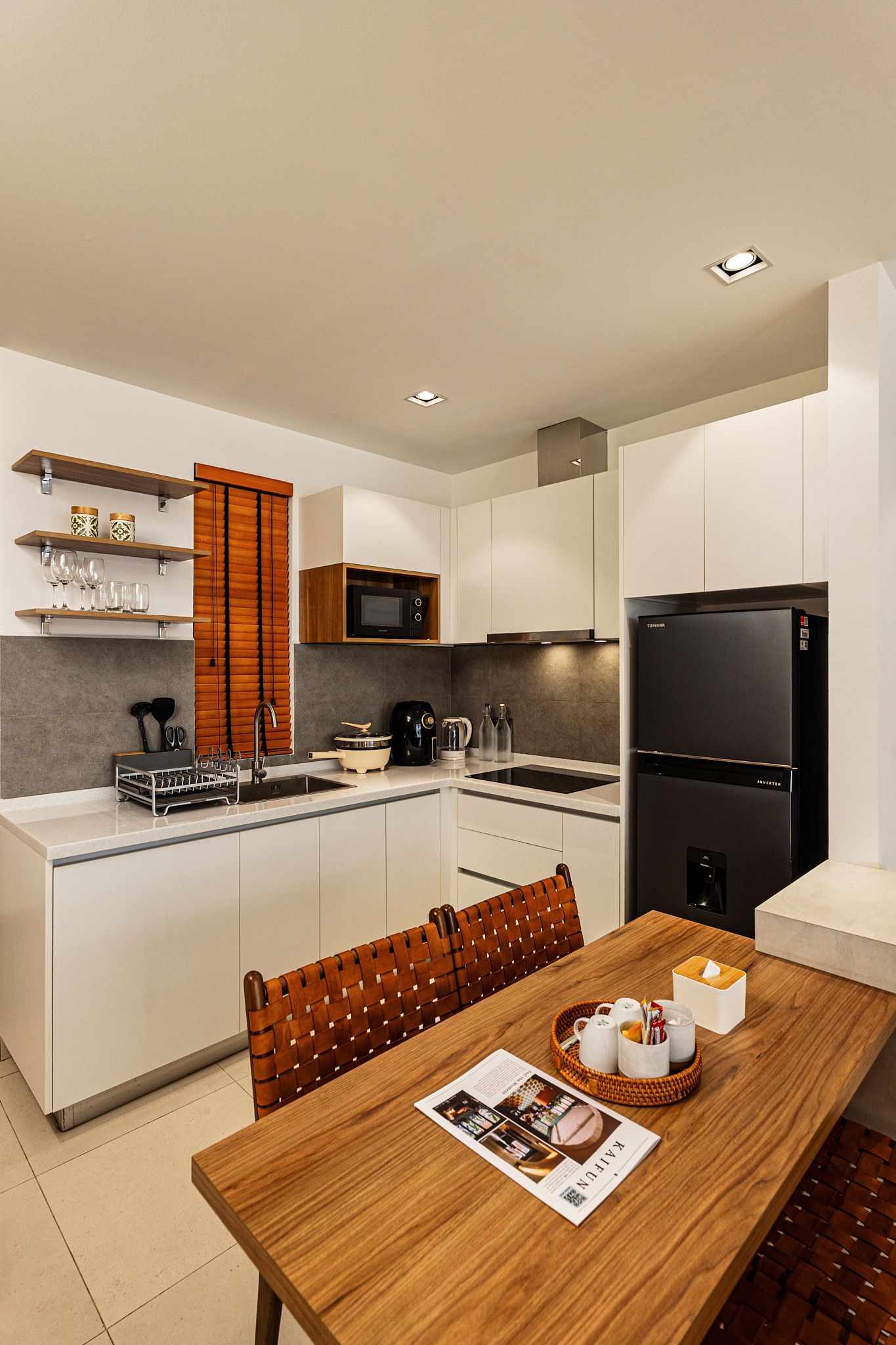 Modern white kitchen with black appliances, wooden dining table, and orange bar stools
