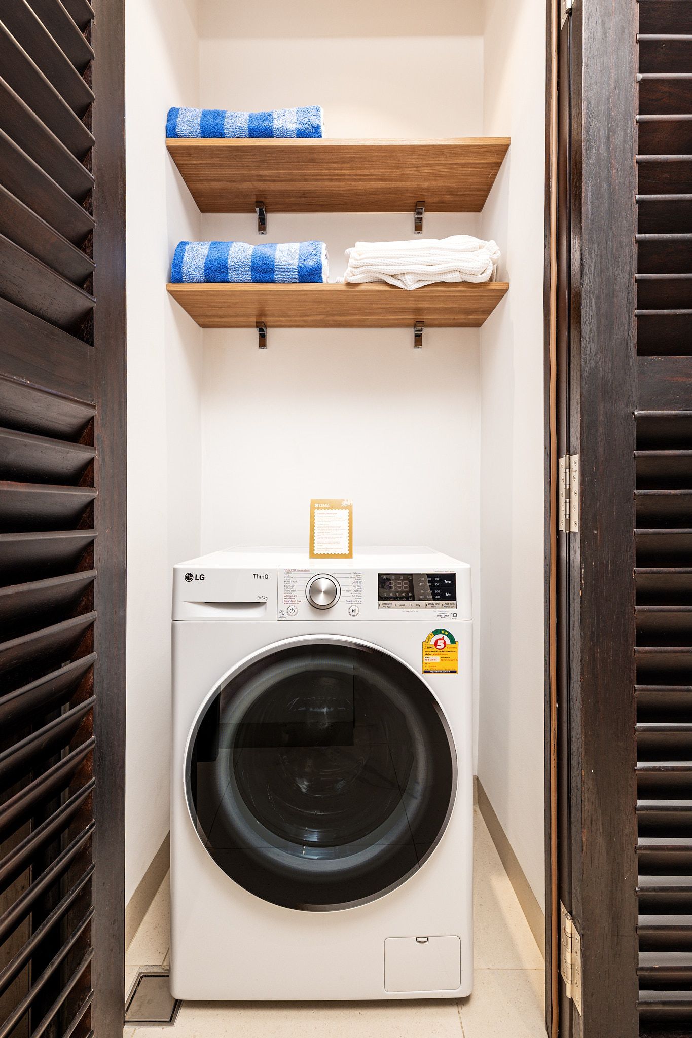 Laundry nook with a front-loading washing machine under wooden shelves holding folded towels and supplies