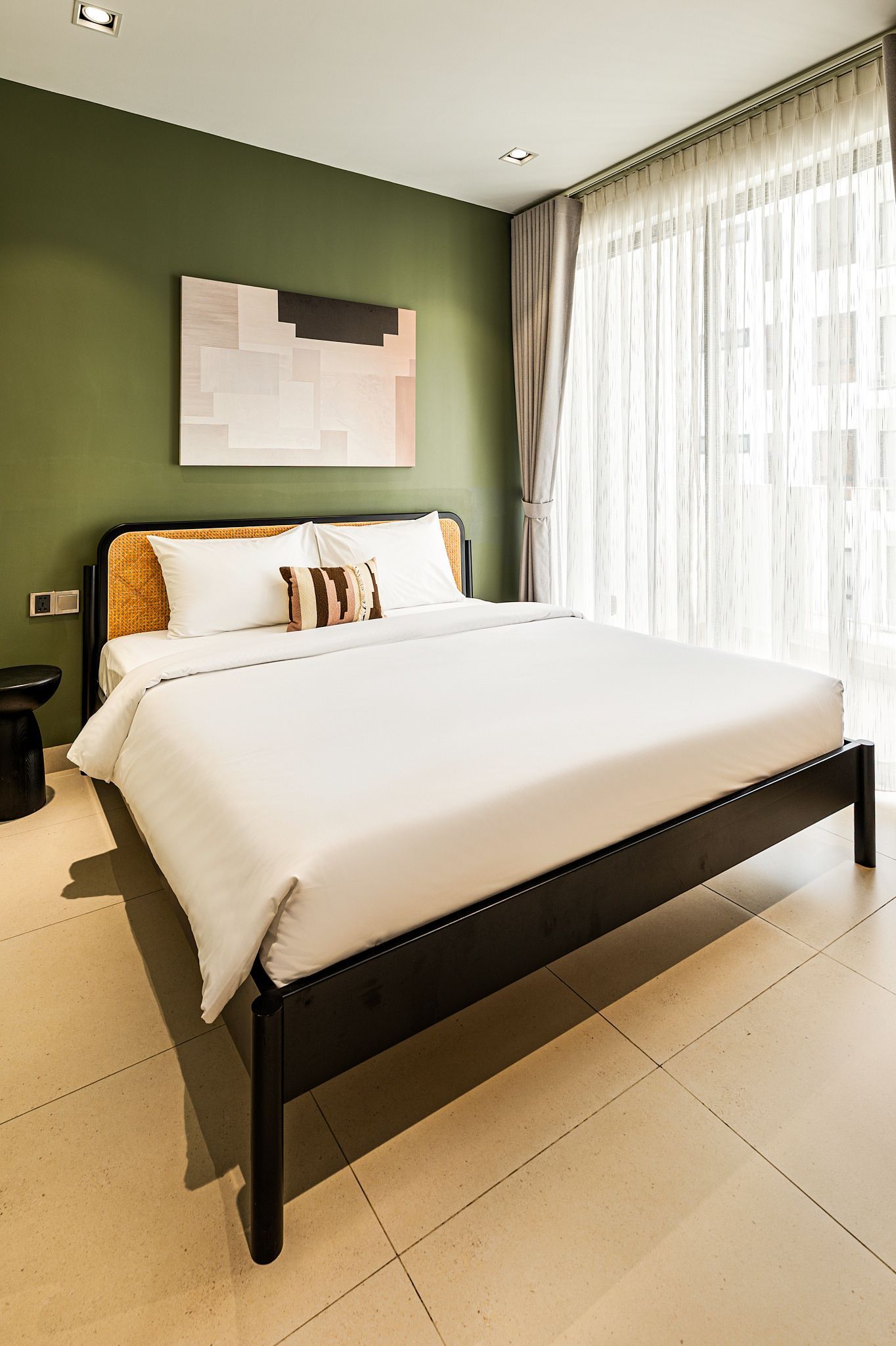Modern bedroom with a neatly made white bed, green accent wall, and sheer curtains letting in sunlight