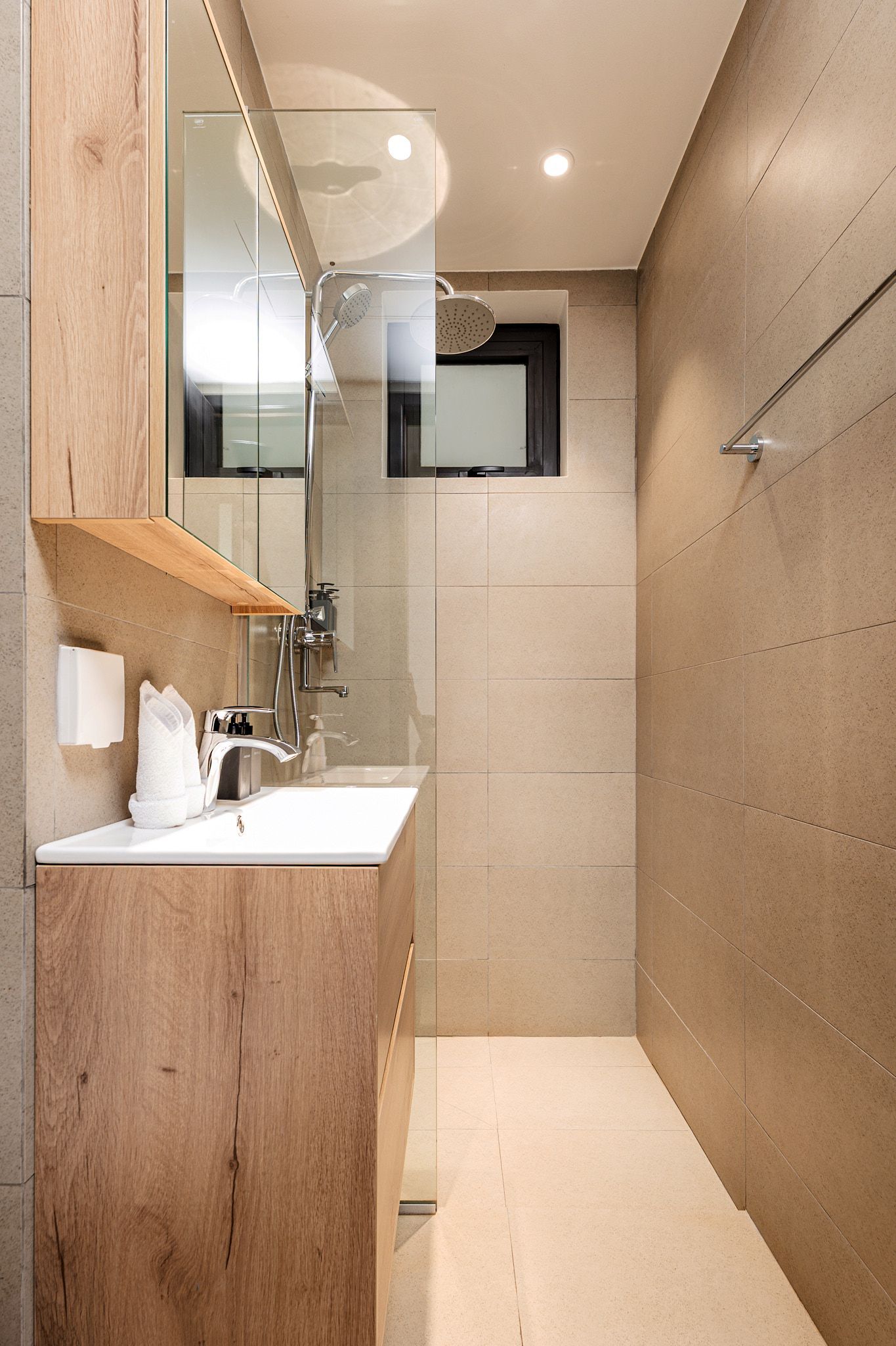 Narrow beige bathroom with glass shower, sink counter, and toilet paper holder beside a window.