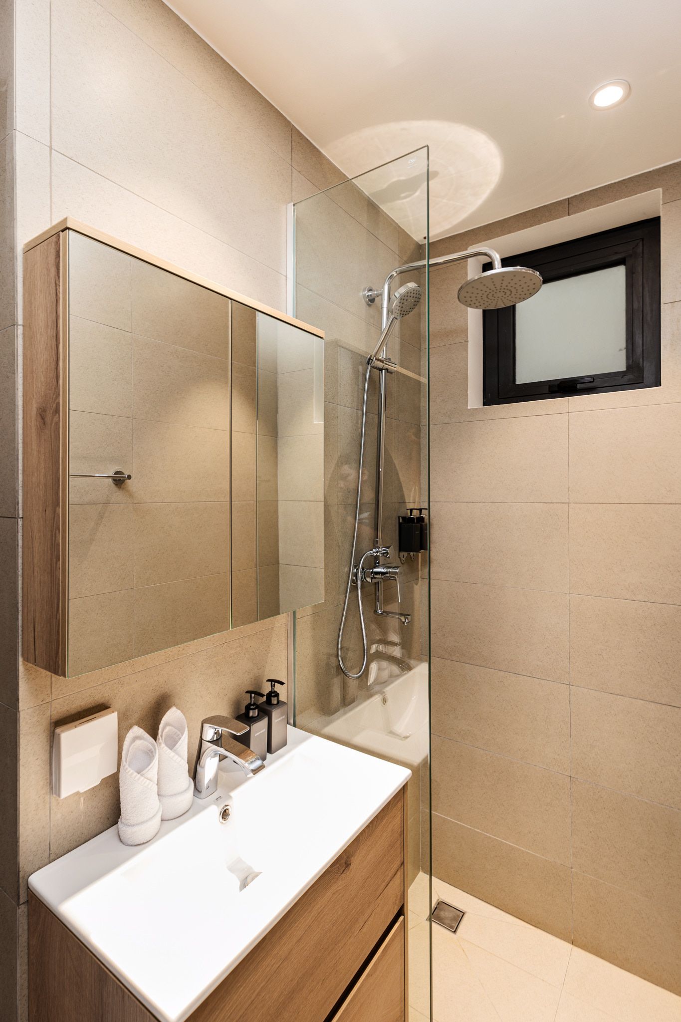 Compact beige bathroom with glass shower, white sink, mirror cabinet, and window above the shower