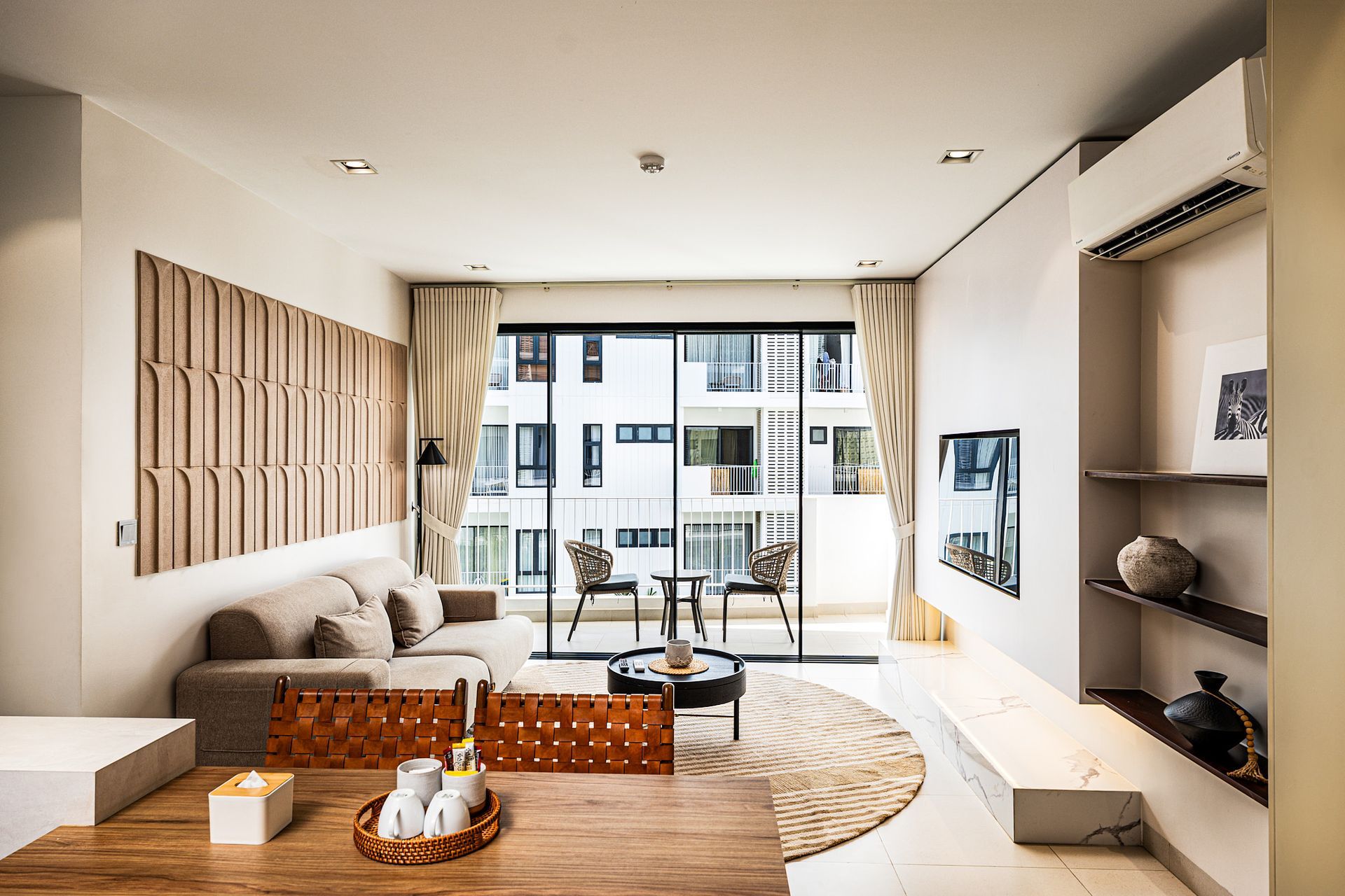 Modern living room with beige sofa, wooden coffee table, shelves, and floor-to-ceiling window to a balcony.