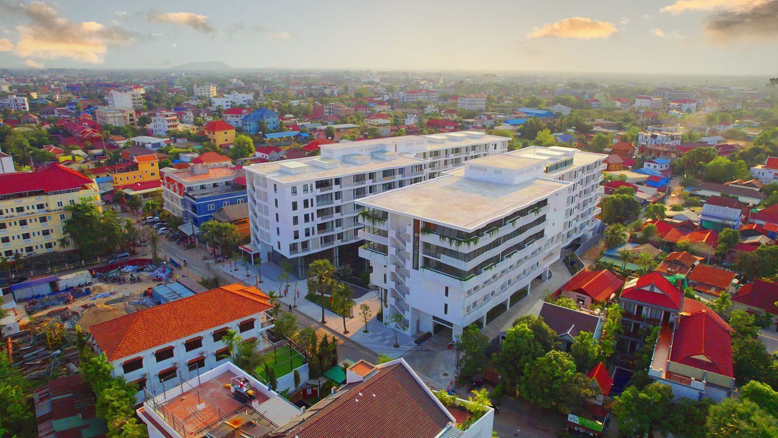 Aerial view of a modern white building with a surrounding cityscape of colorful buildings under a warm sunset.
