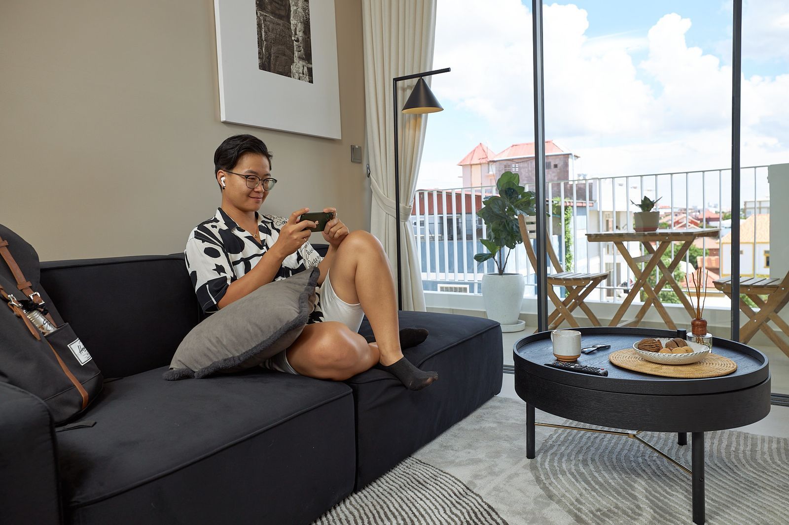 Woman on a black couch plays a game on a phone. Balcony with table/chairs outside a window.