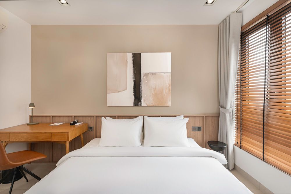 Minimal hotel bedroom with white bed, wooden desk, abstract art, and window blinds