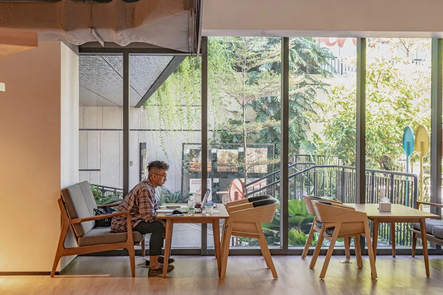 Person working on laptop at a table near a large window overlooking trees. Neutral indoor setting with wooden furniture.