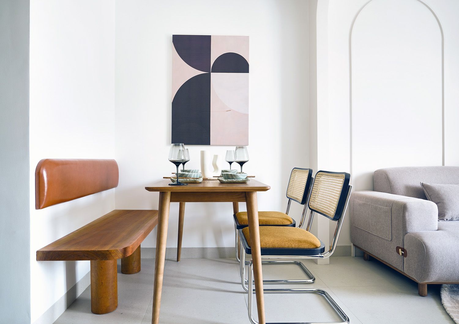 Minimalist dining nook with wooden table, tan bench, woven chairs, and abstract wall art in a bright room