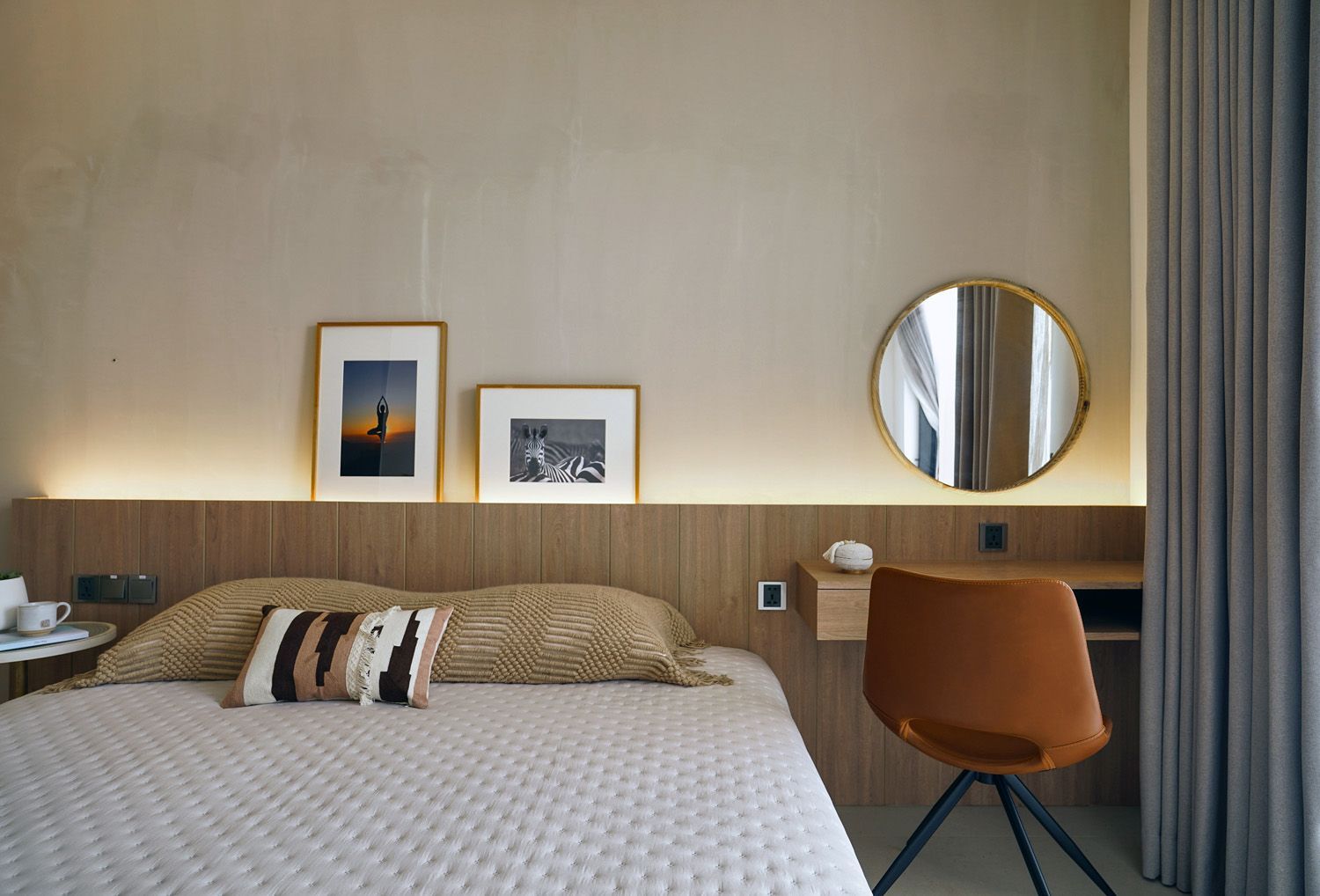 Modern hotel bedroom with bed, framed art, round mirror, and brown chair beside a curtained window