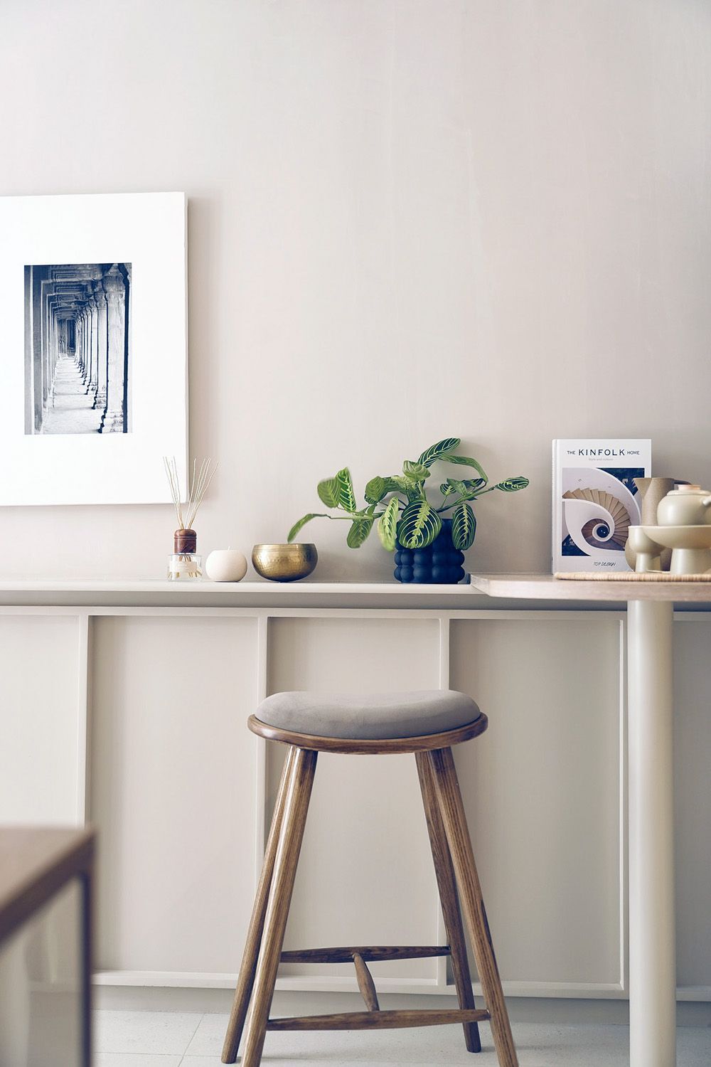 Minimalist desk with a stool, vase of flowers, framed art, and decor against a beige wall.