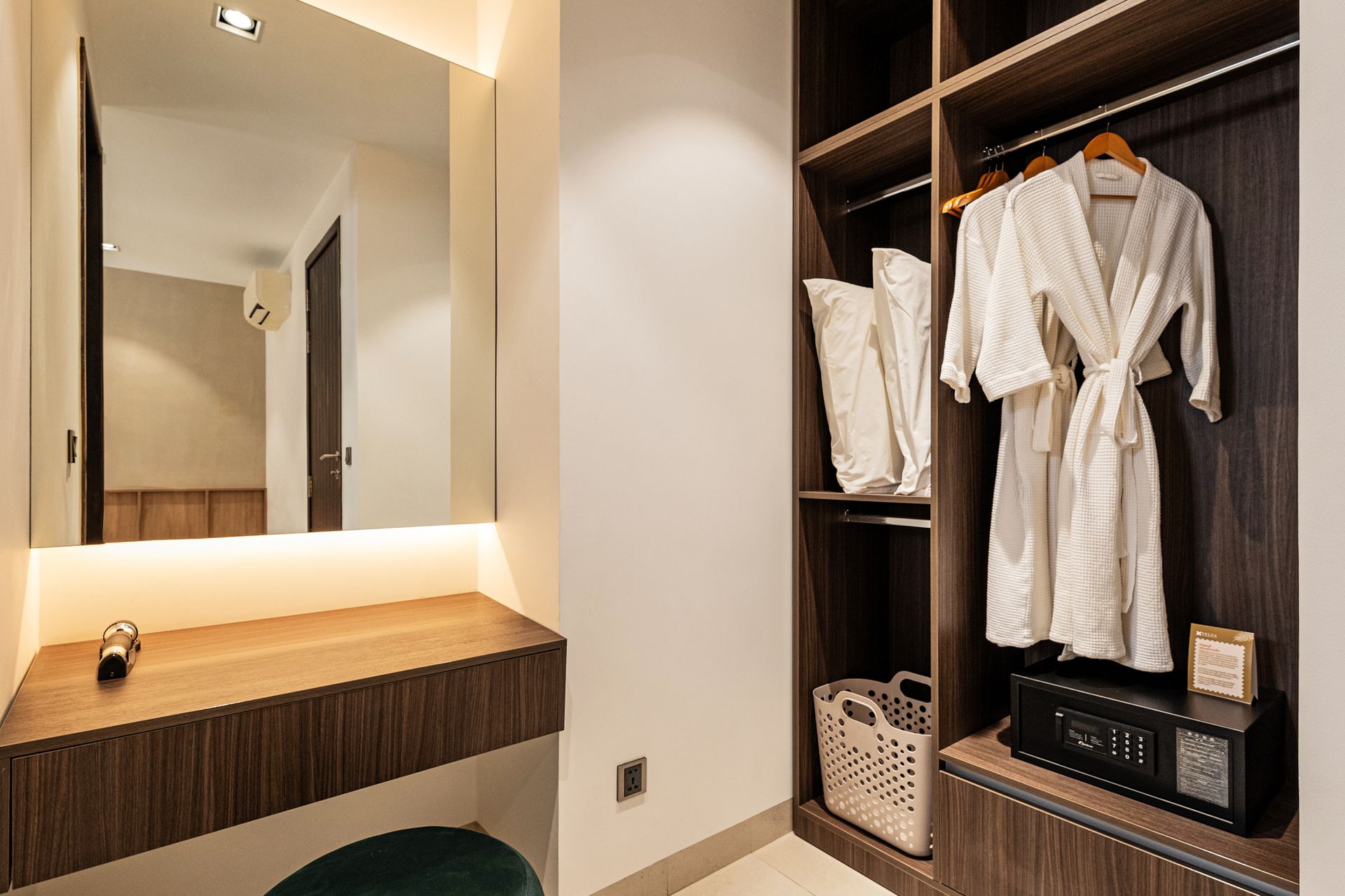 Modern hotel dressing area with robe, mirror, wooden vanity, and open closet with folded towels and luggage rack