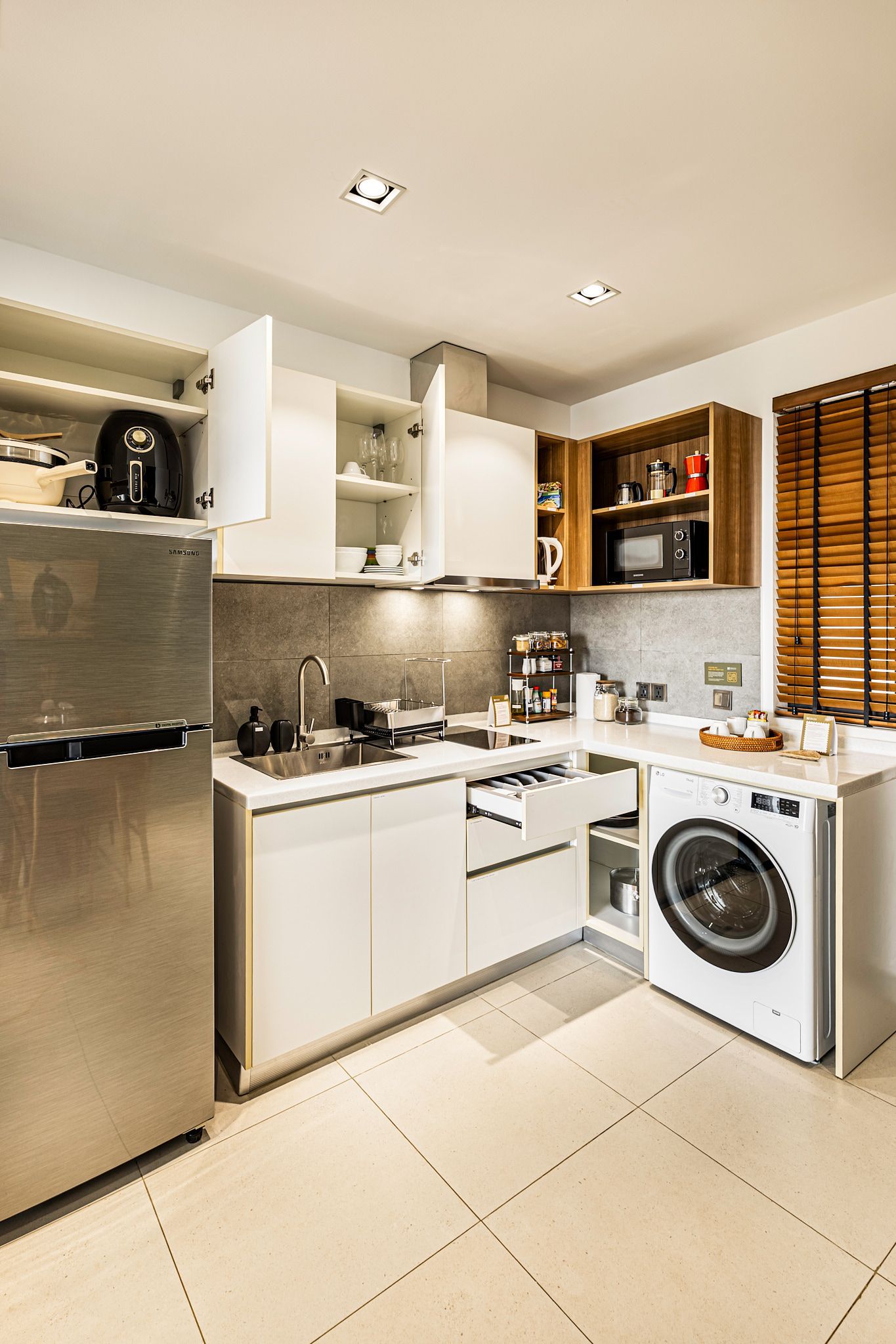 Compact modern kitchen with stainless fridge, white cabinets, stove, and washing machine by a window with blinds
