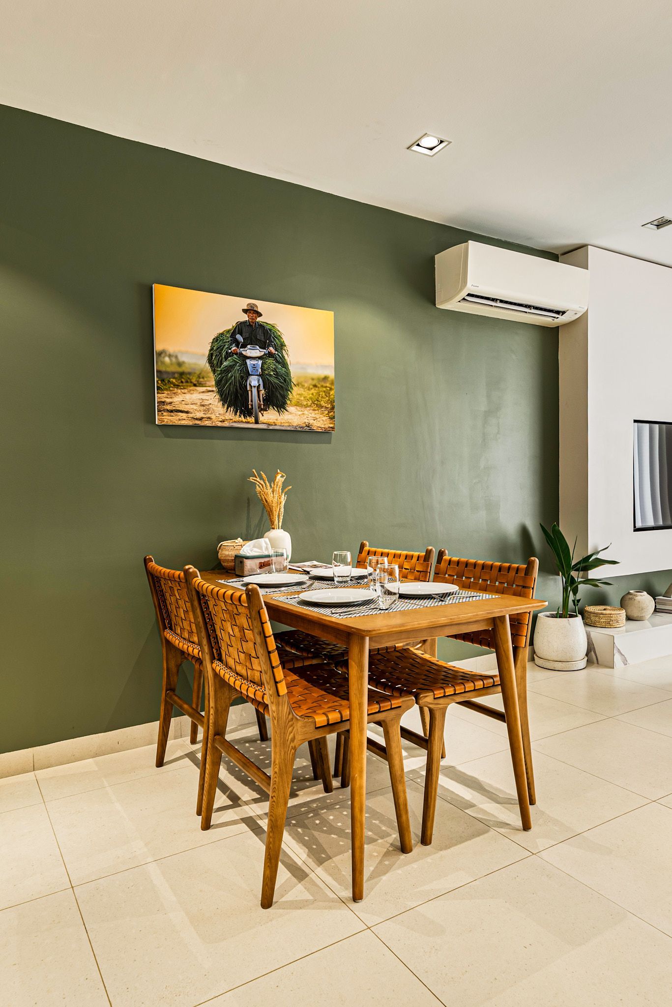 Dining table with woven chairs in a modern room, olive green accent wall, framed art, and potted plants.