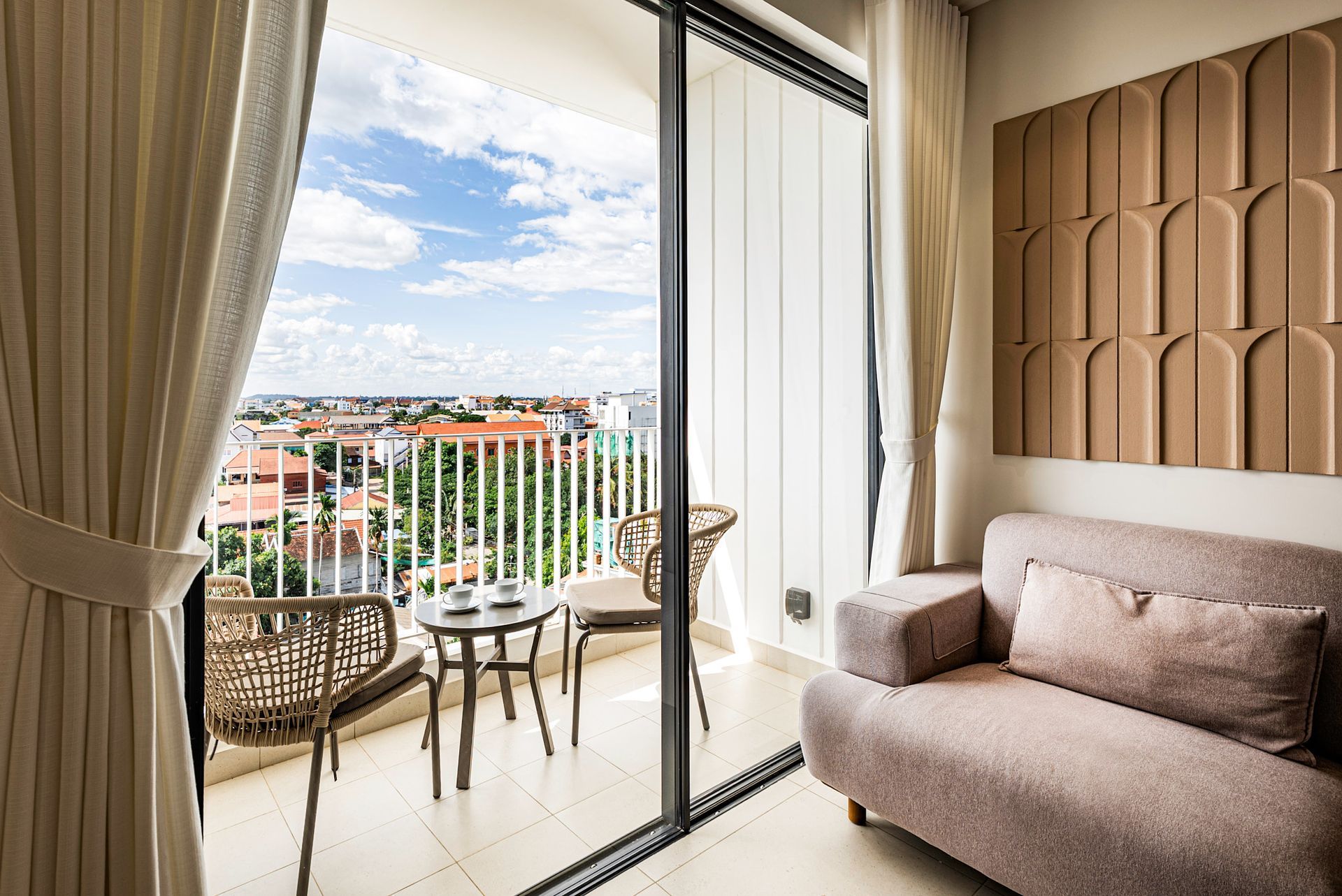 Bright balcony view through sliding doors beside a beige sofa and table, with city buildings and blue sky.