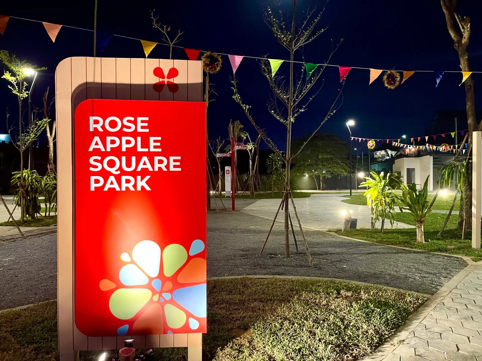 Sign for Rose Apple Square Park at night, red with logo. Park pathway, colorful flags.