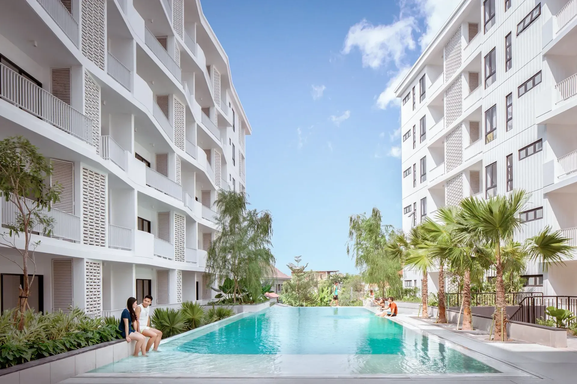 Swimming pool between white buildings. Couple sits on edge. Sunny day.