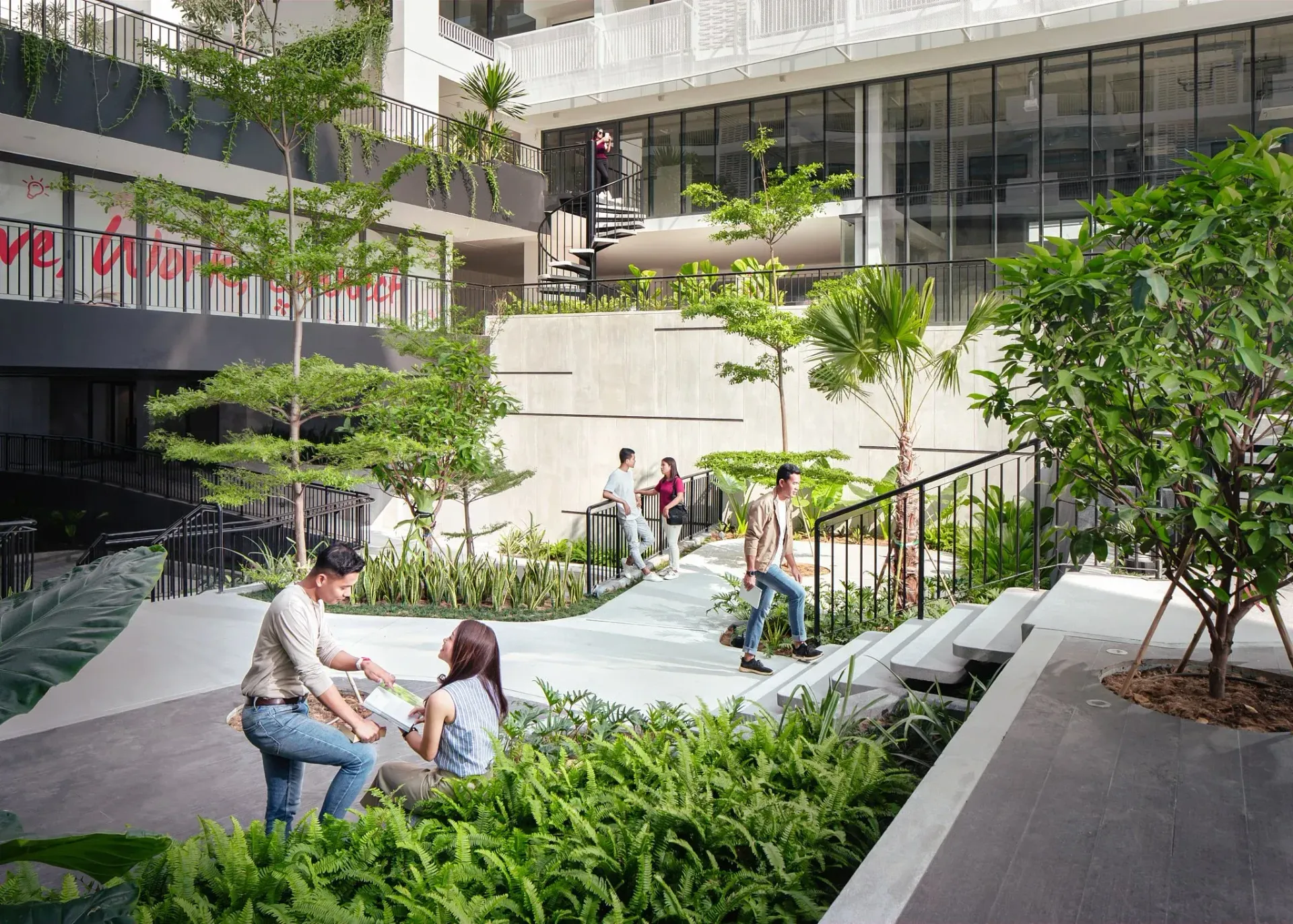 People sitting in a modern courtyard garden with trees, steps, and glass buildings around them