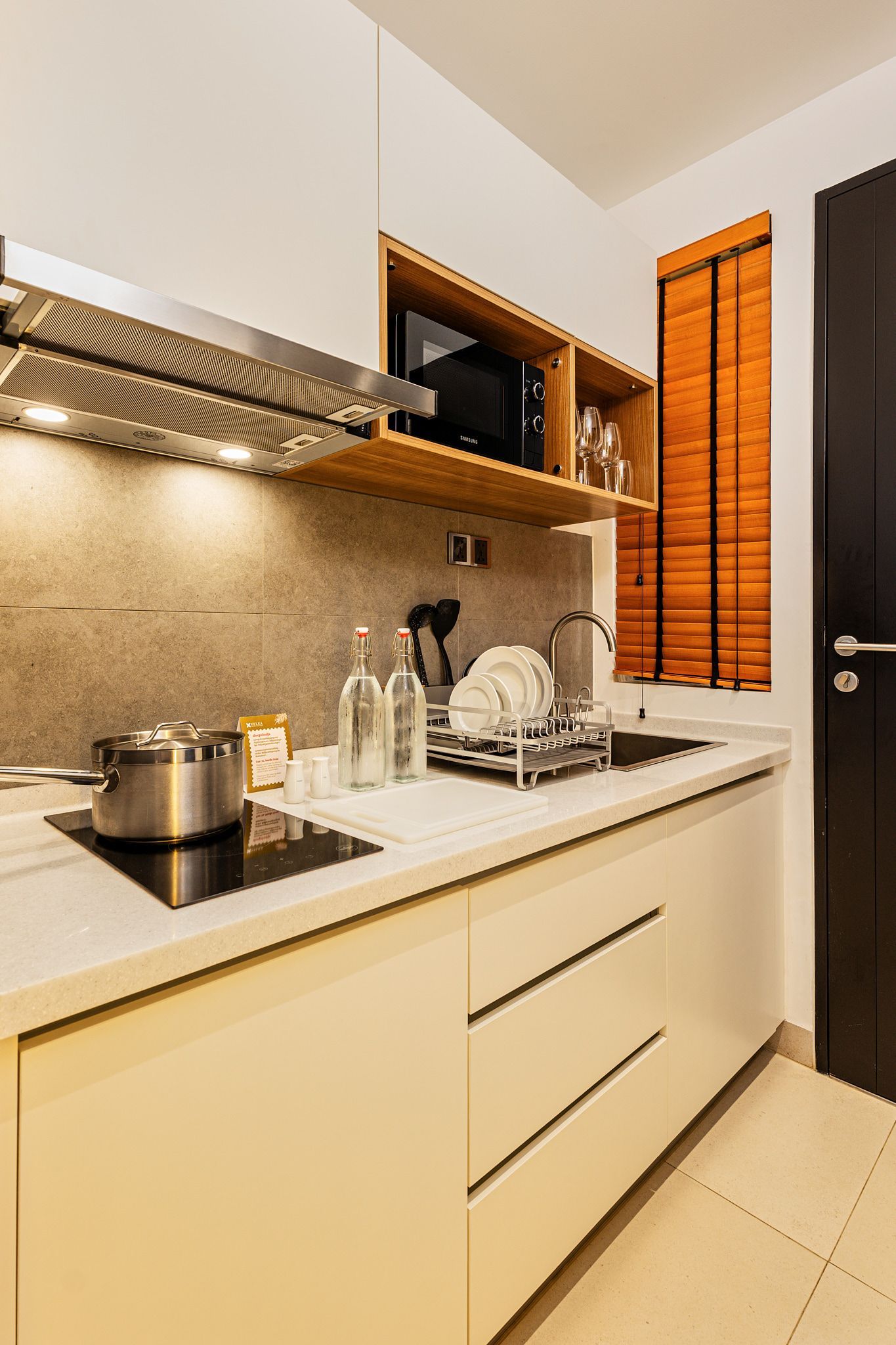 Compact modern kitchen with stovetop, pots, white cabinets, sink, and warm window blinds