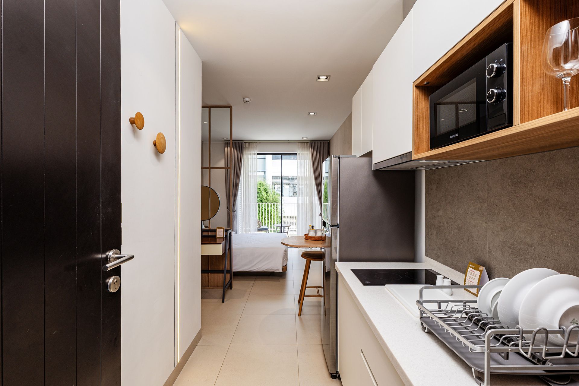 Narrow modern kitchen corridor with dark cabinets, white counters, and a bright windowed dining area beyond