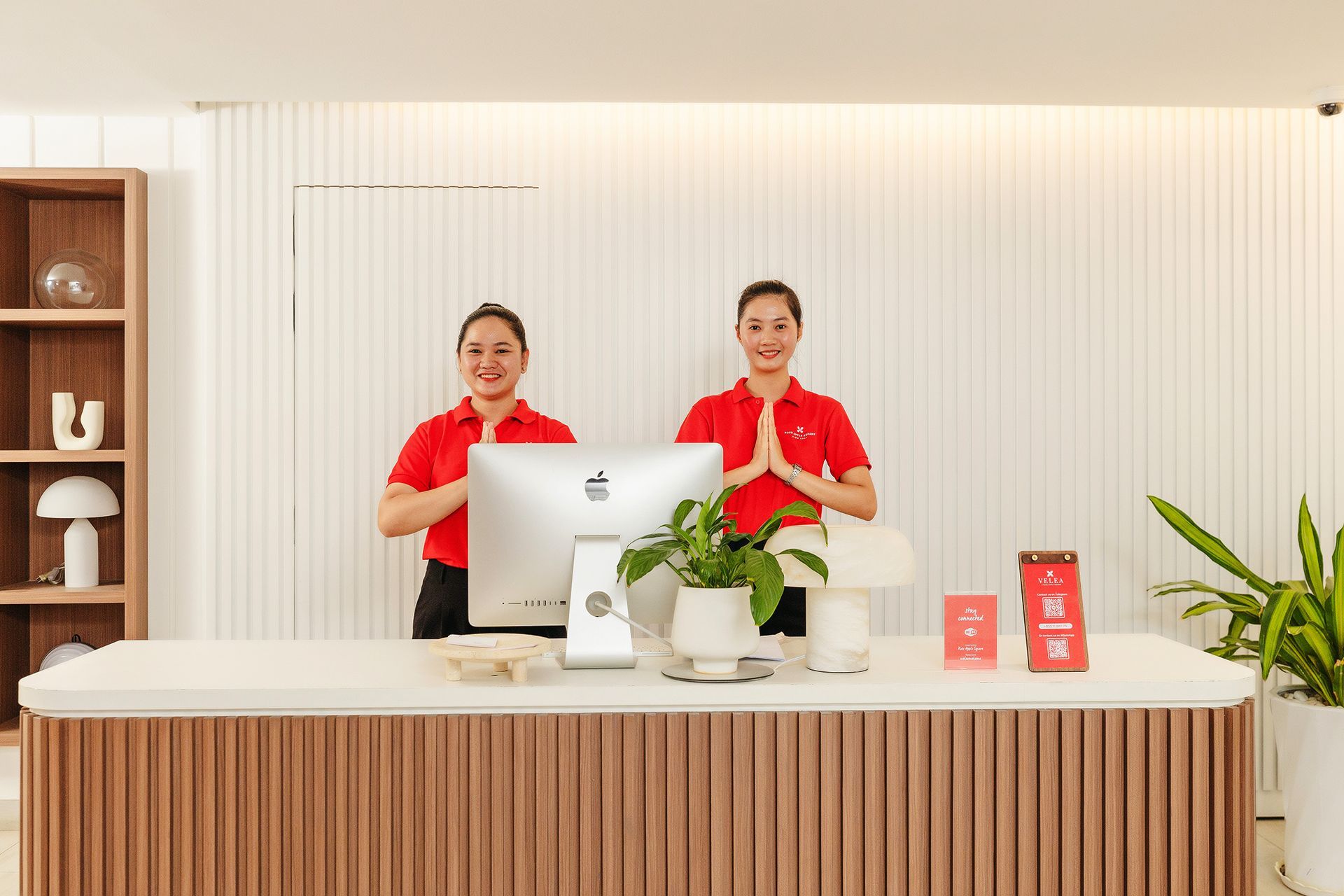 Two men at a hotel reception desk, one handing a card to a staff member. Modern setting with a laptop and shelves.