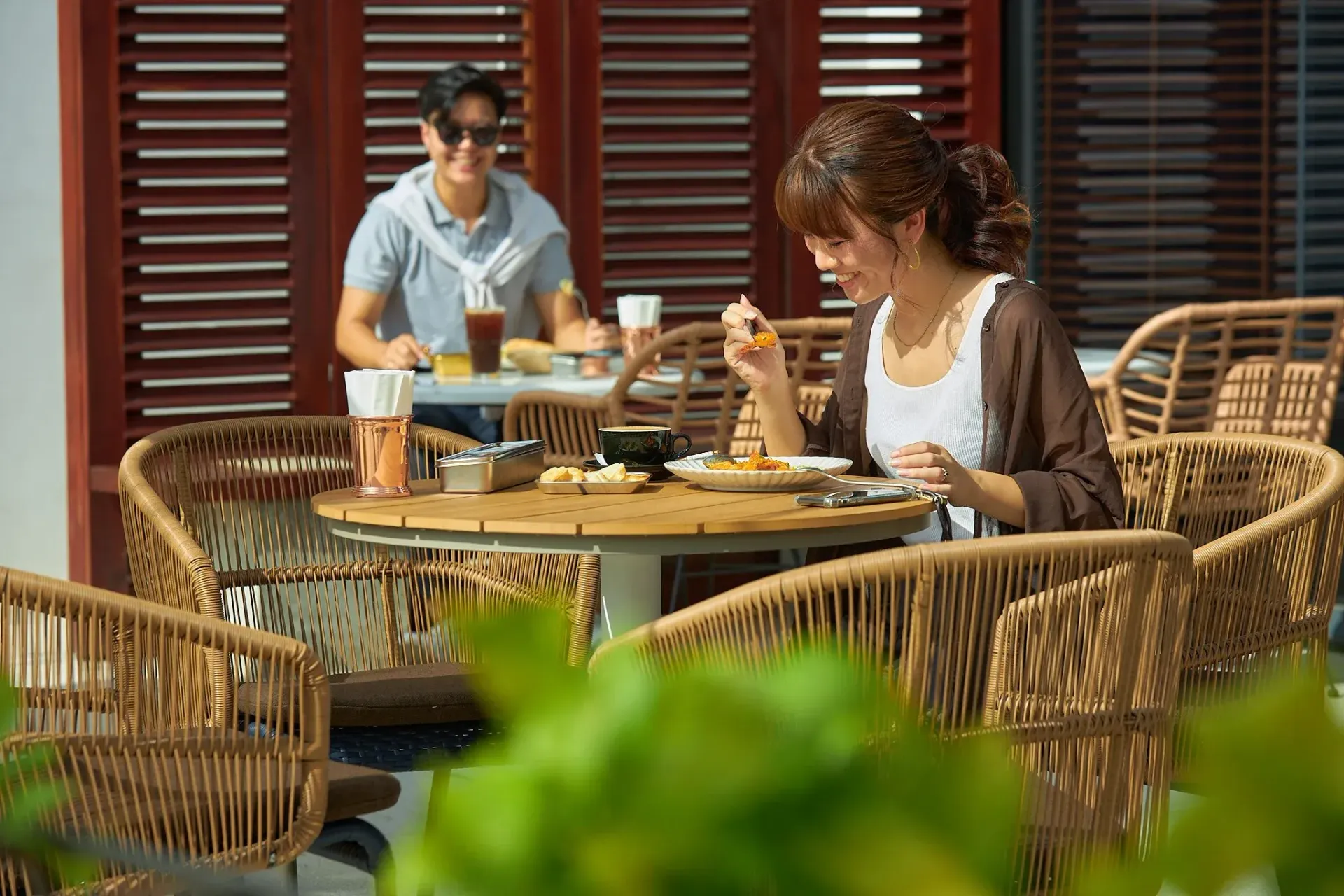 Couple eating at a restaurant with wicker chairs and red shutters. Woman eating, man drinking.