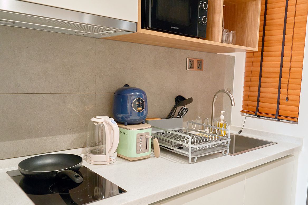 Compact kitchen counter with sink, dish rack, kettle, and blue appliance beside a window with blinds