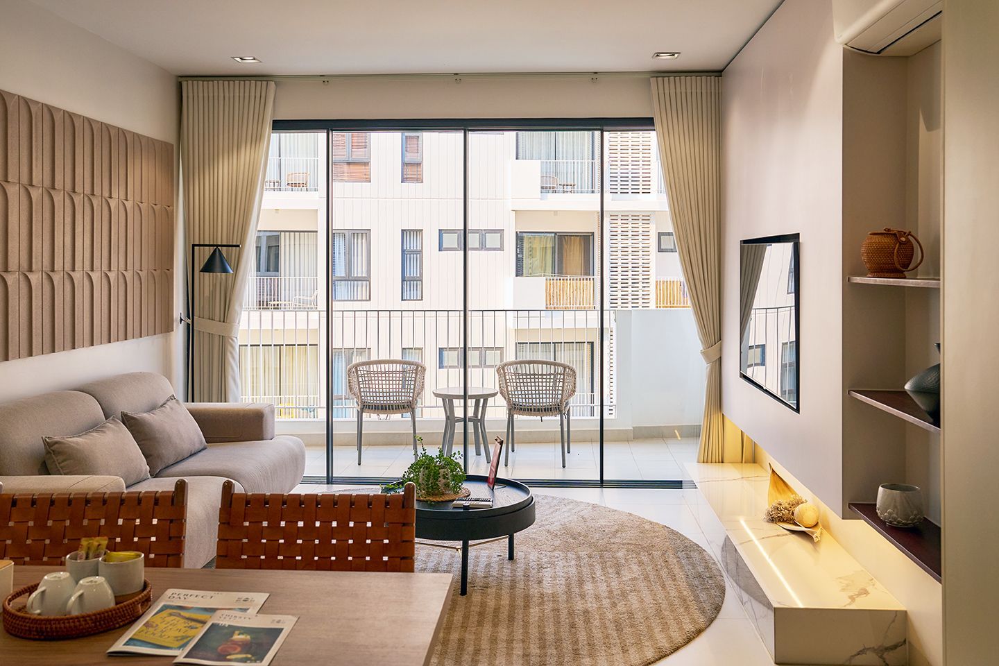 A modern, minimalist apartment living room with a light gray sofa, wooden coffee table, dining area, and white kitchen.