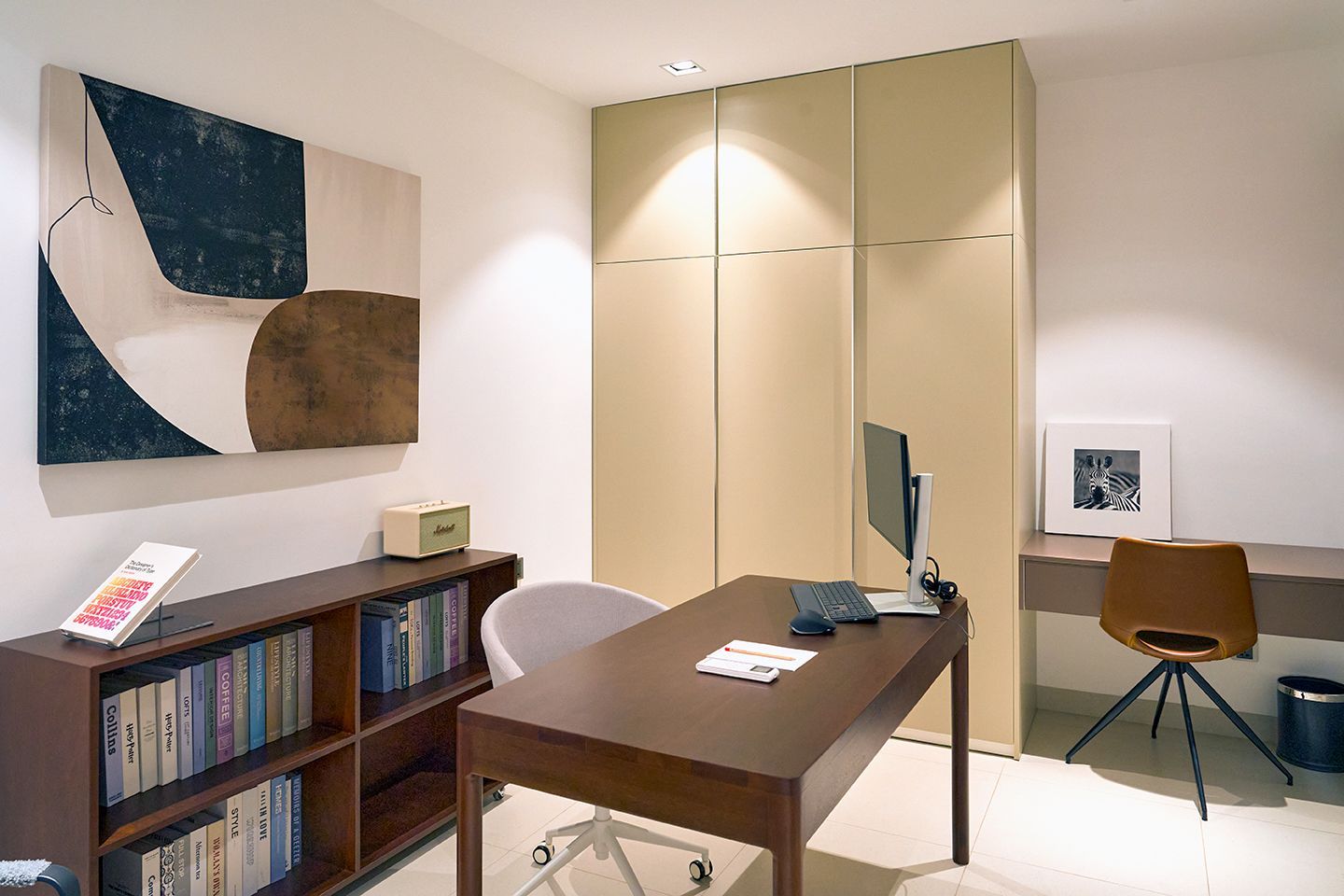 Small office with desk, bookshelf, abstract wall art, and beige cabinets in a minimalist room.
