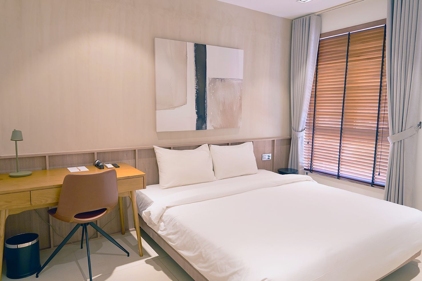 Bright bedroom with a neatly made bed, desk, chair, wall art, and window blinds.