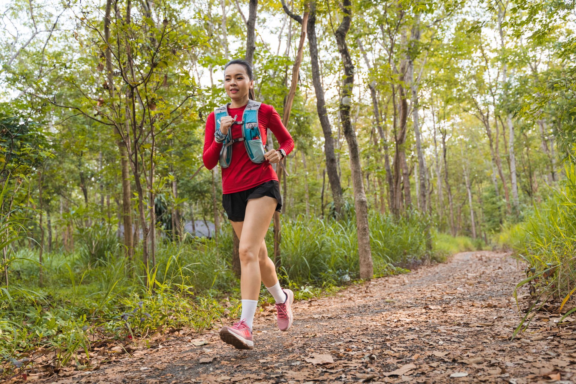 Trail runner in a red jacket jogging through a wooded forest path with a blue hydration pack
