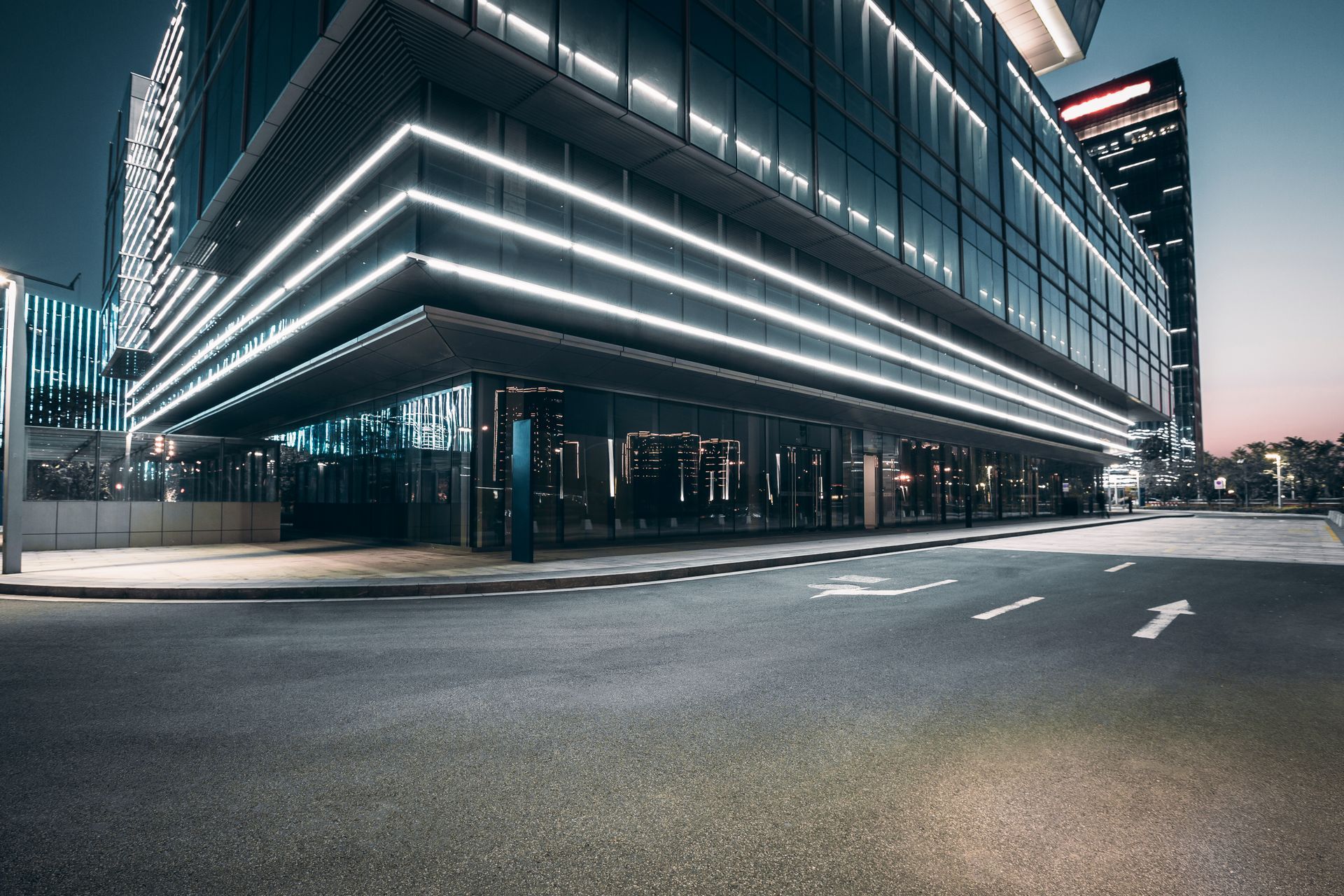 Modern glass office building at dusk, with illuminated edges and an empty street in front.