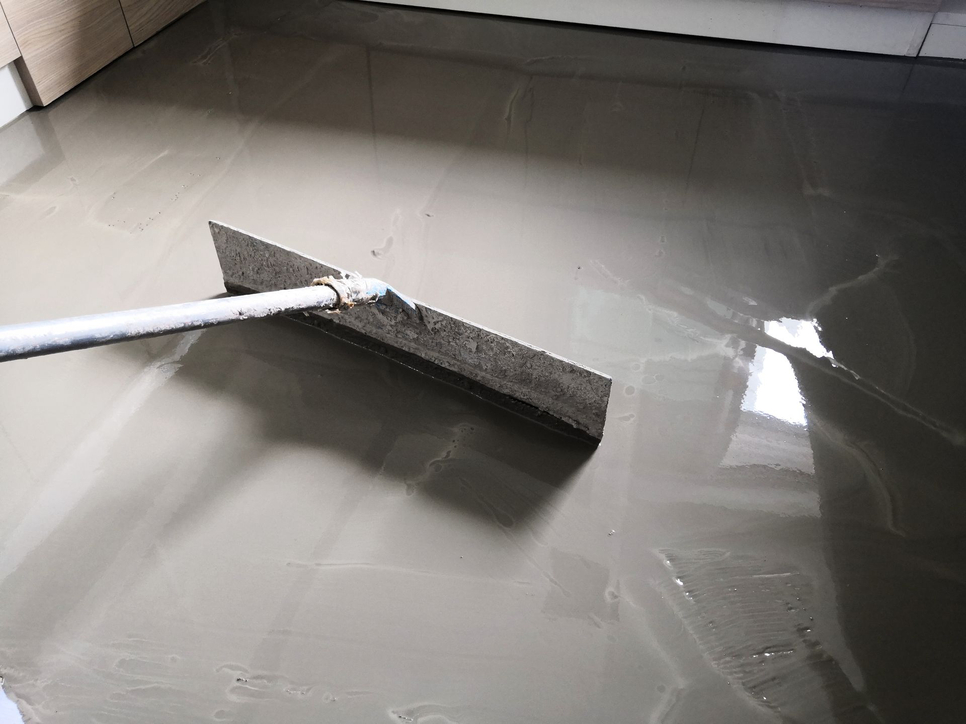 Squeegee spreading gray liquid across a glossy floor near a baseboard board