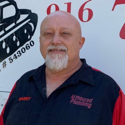 Smiling man in navy work shirt with “Armoured Plumbing” logo standing before a service truck.
