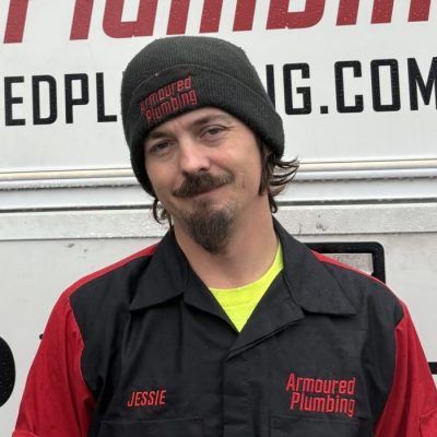 Man in black and red work shirt and beanie, standing in front of a sign, smiling slightly.