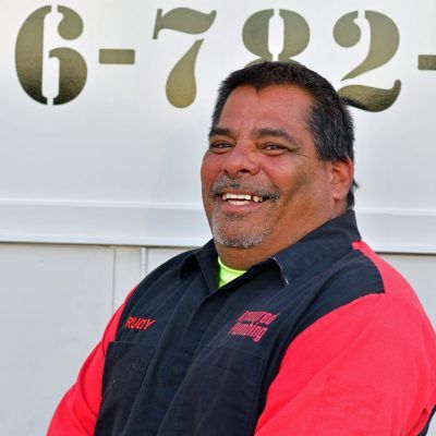 Smiling man in a black and red jacket standing in front of a white wall with the number 672.