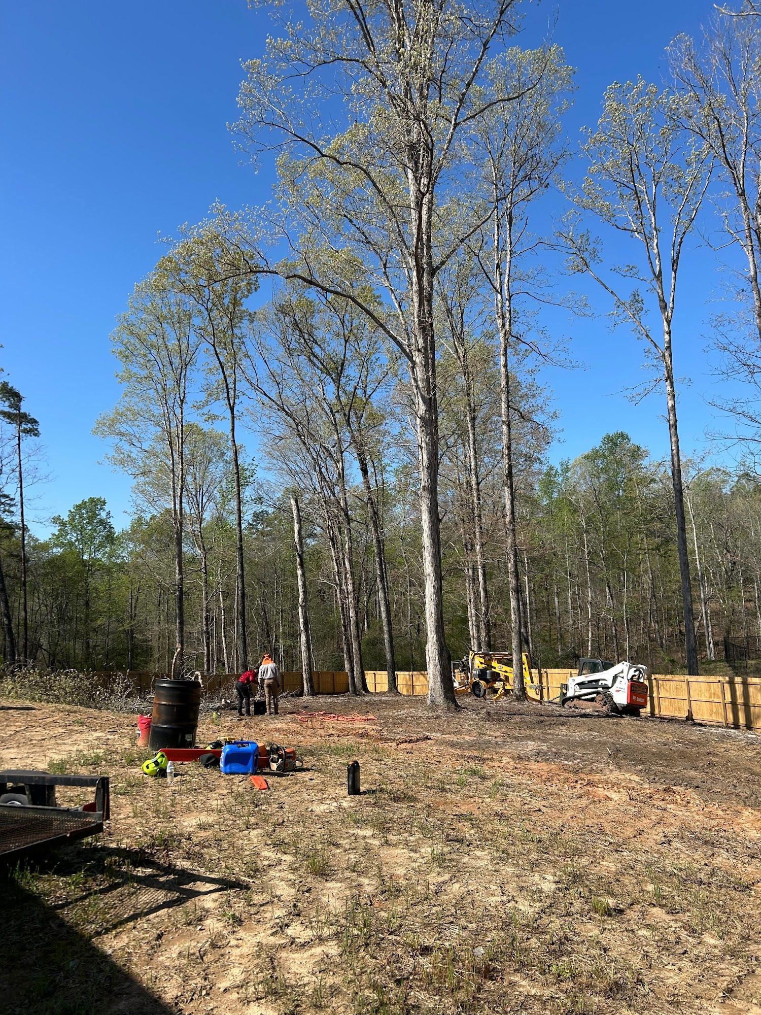 Construction site with trees, blue sky, and two workers near a wooden fence and machinery.