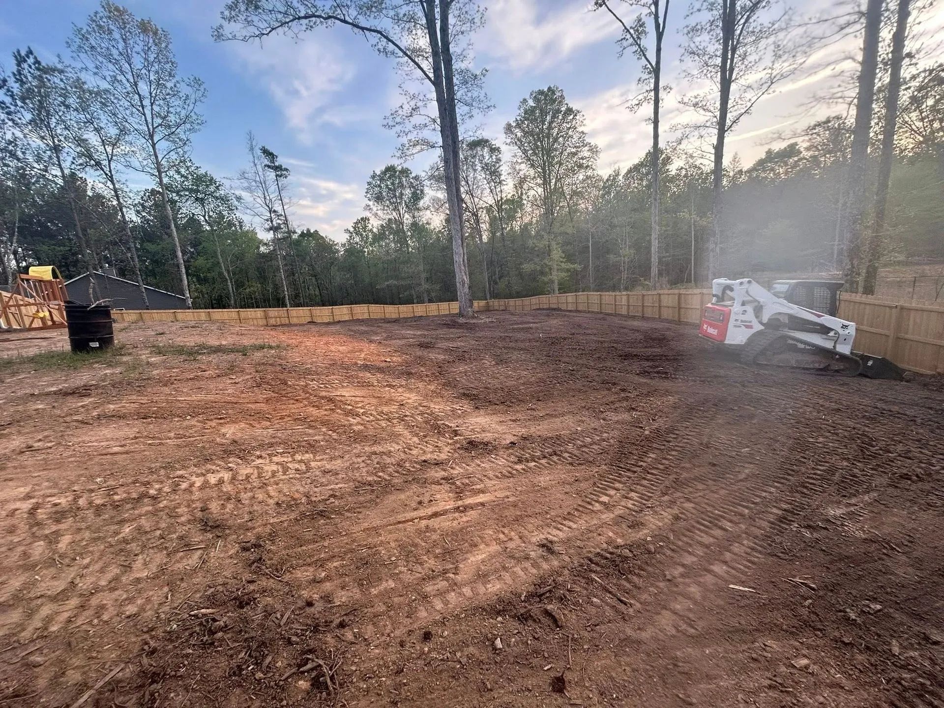 Cleared dirt lot with trees at sunset; skid-steer works on the right beside a black barrel on the left