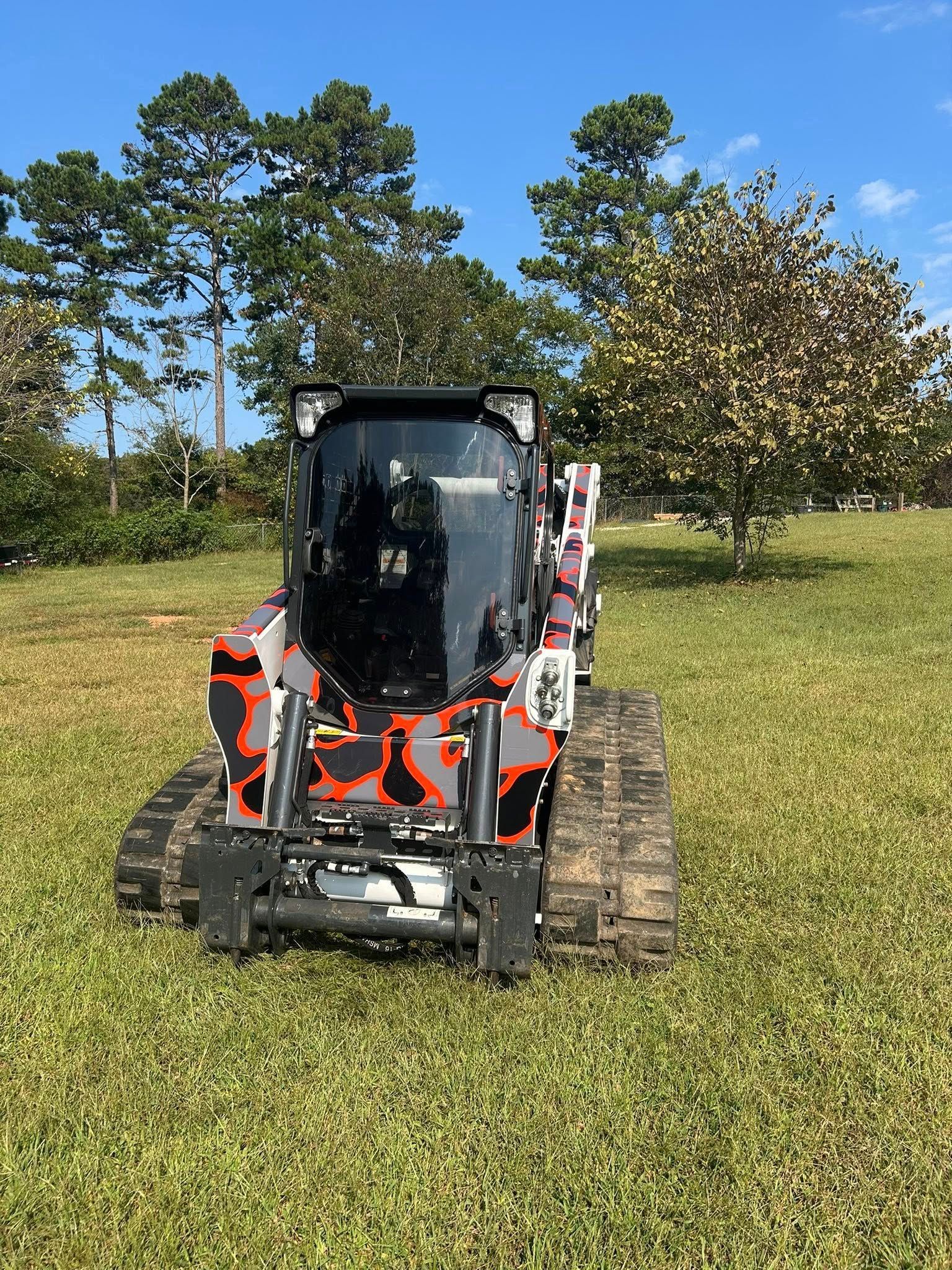 A small, camouflaged skid steer tractor on tracks sits in a grassy field with trees in the background.