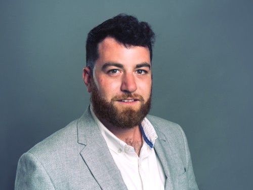 Portrait of a bearded man in a light gray blazer and white shirt against a gray background