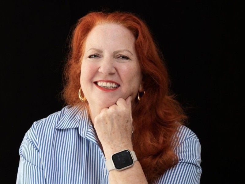 Smiling red-haired person in a striped shirt posing against a black background, chin resting on hand.
