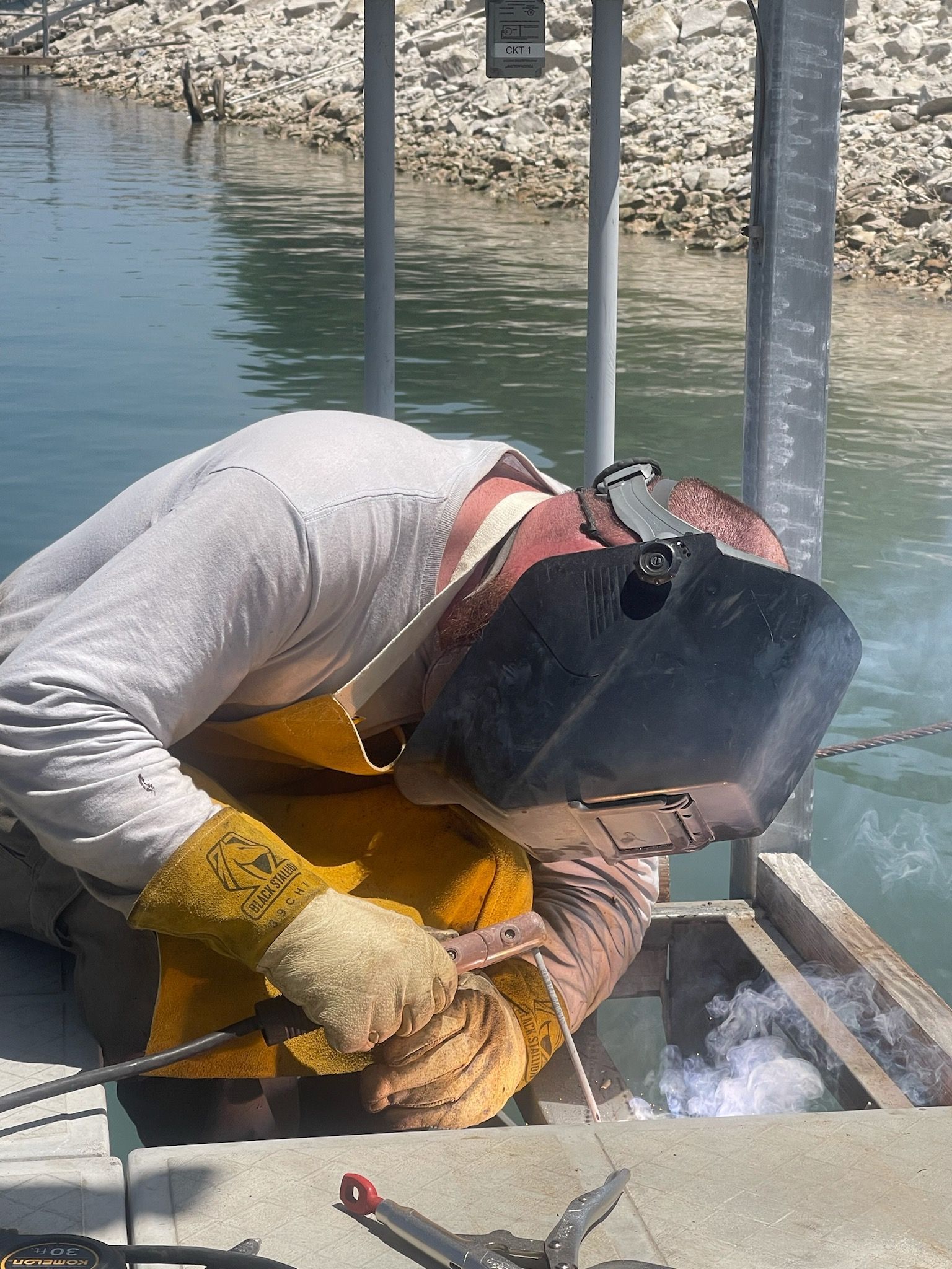 Welder in protective gear welding metal on a dock by water. Sparks and smoke visible.