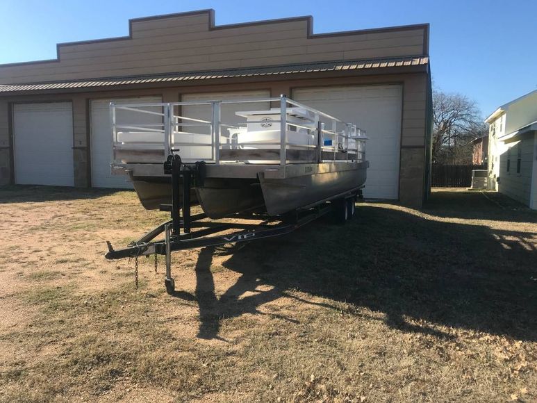 Pontoon boat on a trailer in front of a tan building with garage doors on a sunny day.